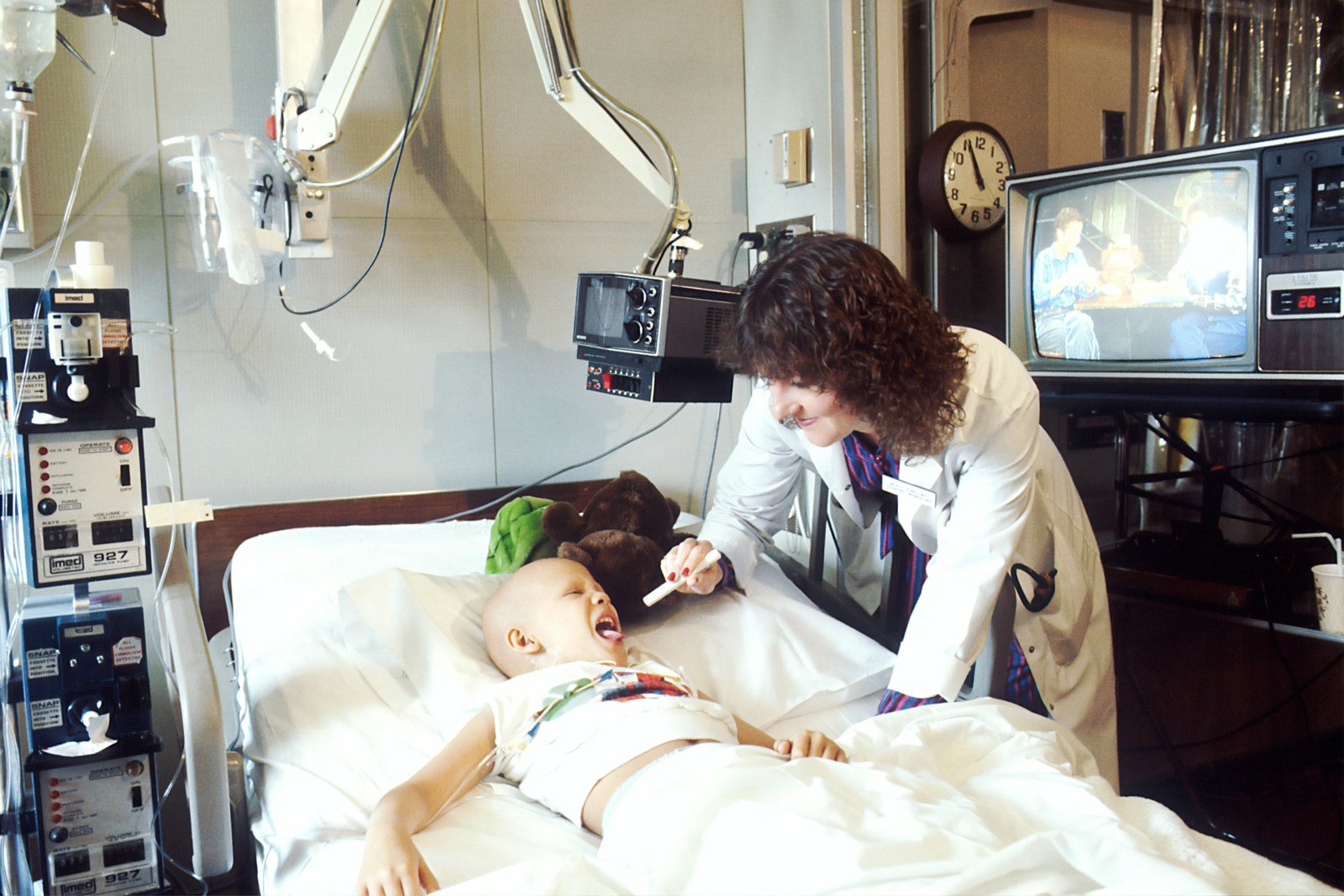 A medical professional in a white coat performs an ultrasound examination on a young child lying on a hospital bed, with cardiac monitoring equipment and display screens visible in the background.