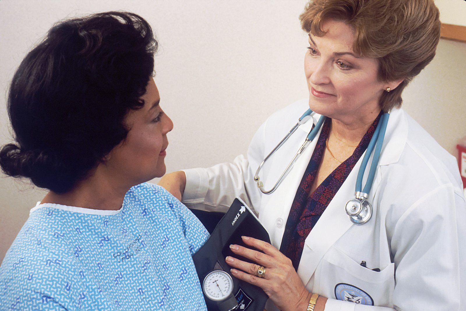 A female healthcare provider in a white coat with a stethoscope examines a patient wearing a light blue hospital gown, performing a medical consultation in a clinical examination room.
