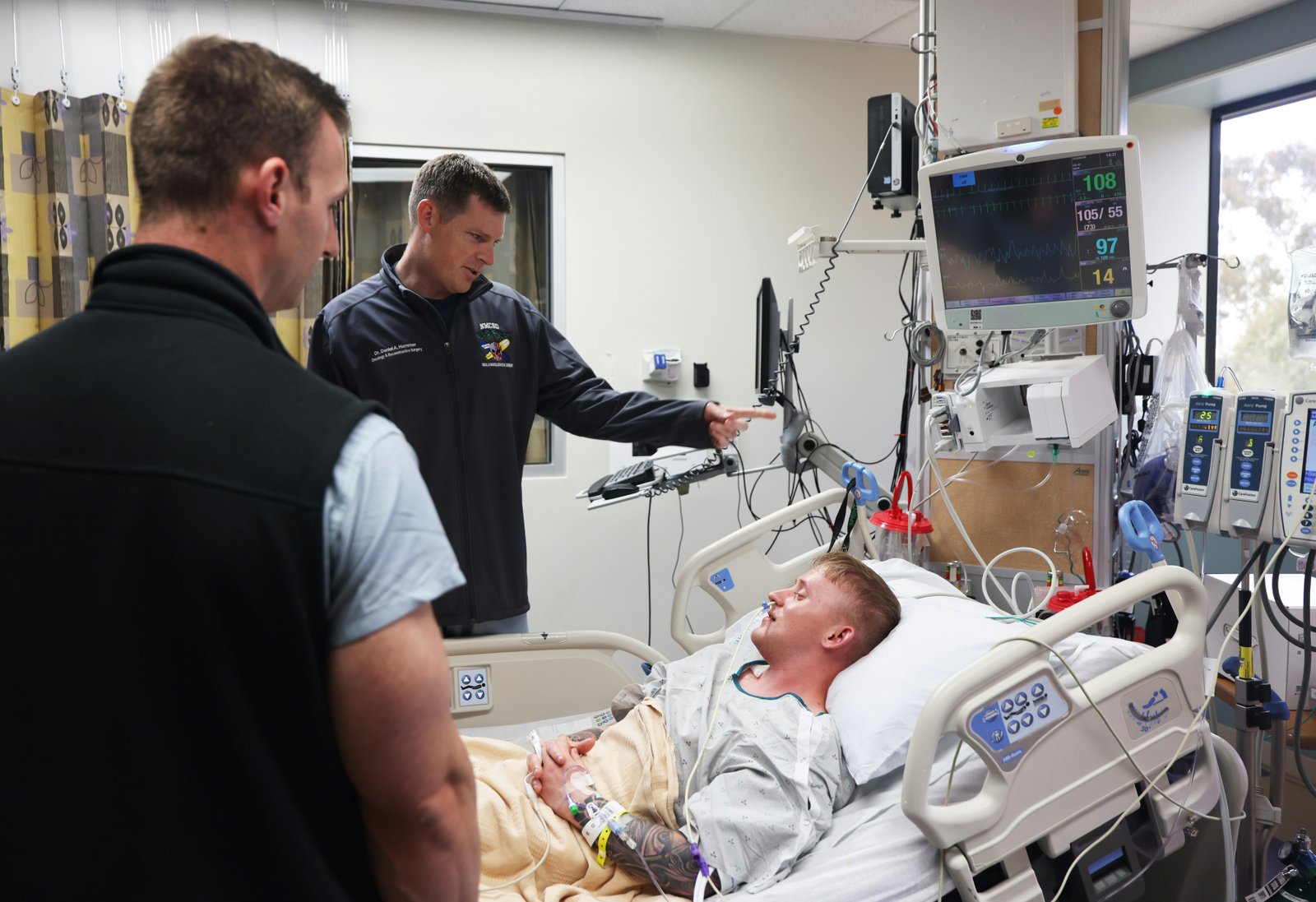 A patient lying in a hospital ICU bed wearing medical monitoring equipment while two healthcare workers in dark clothing stand beside the bed observing medical monitors and equipment in a clinical hospital room.
