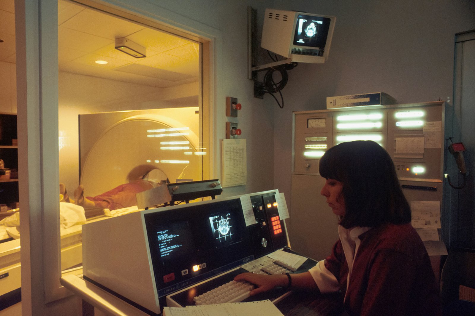 A person in dark clothing sits at a desk operating a computer with a display monitor showing what appears to be medical or data management software, with a telephone visible on the desk, in a professional office setting with glass-walled rooms visible in the background.