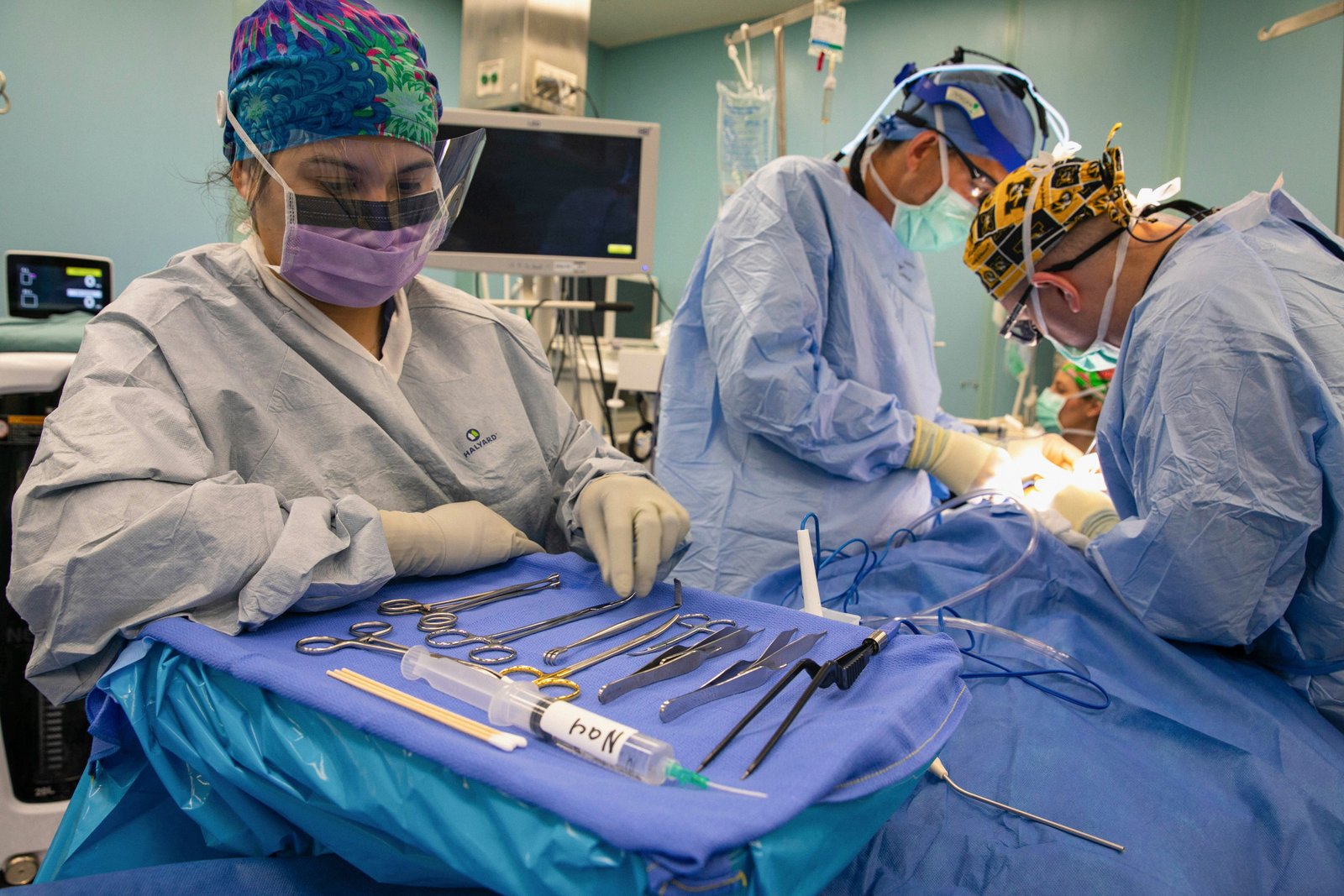 Three surgical team members in sterile protective gear and surgical masks perform a procedure on a blue drape-covered operating table, with surgical instruments visible on a tray in the foreground and medical equipment visible in the background operating room.