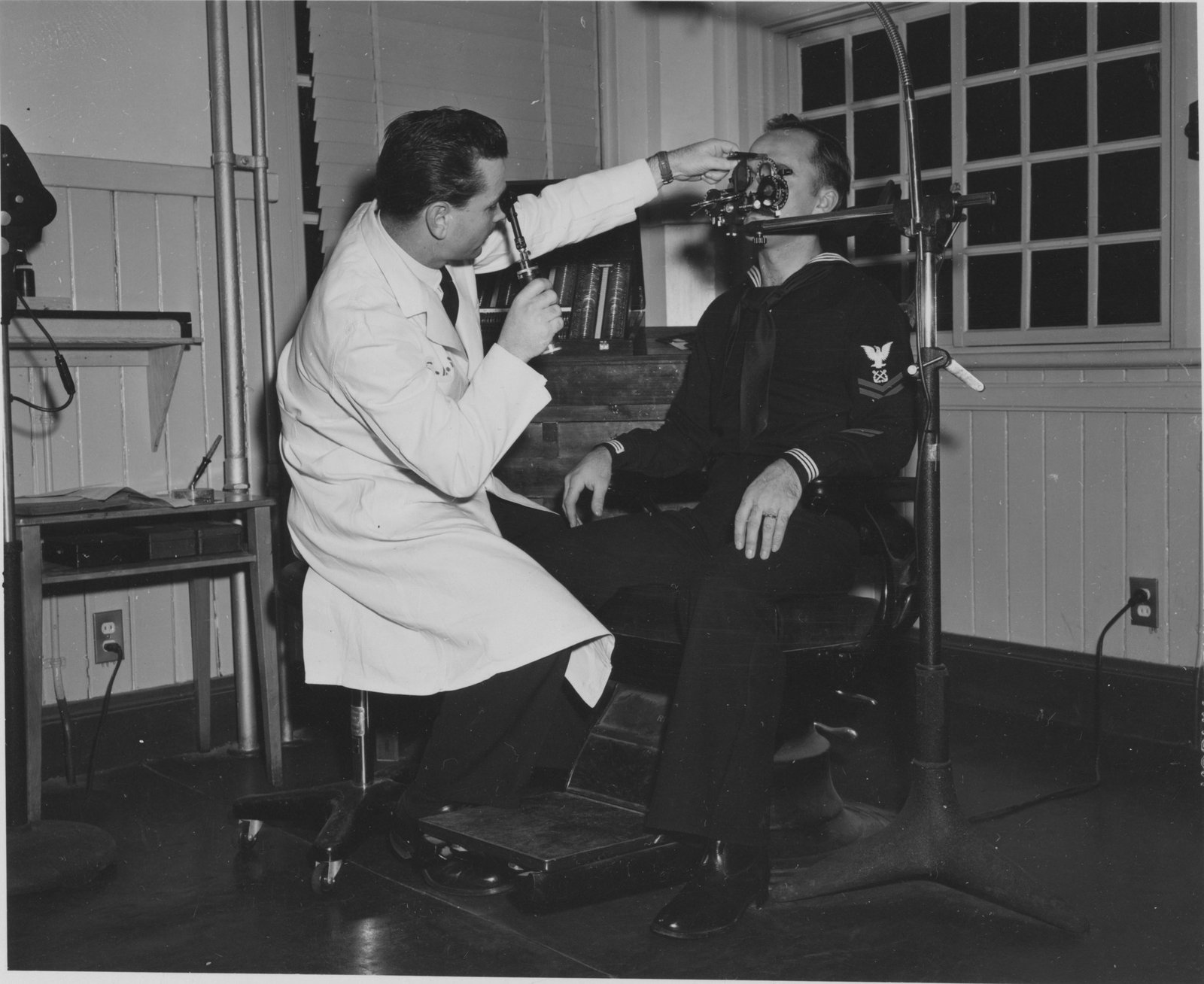 A black and white photograph showing a man in a light-colored shirt examining or adjusting medical/laboratory equipment while another person in dark clothing sits nearby observing, with laboratory benches and gridded windows visible in the background.