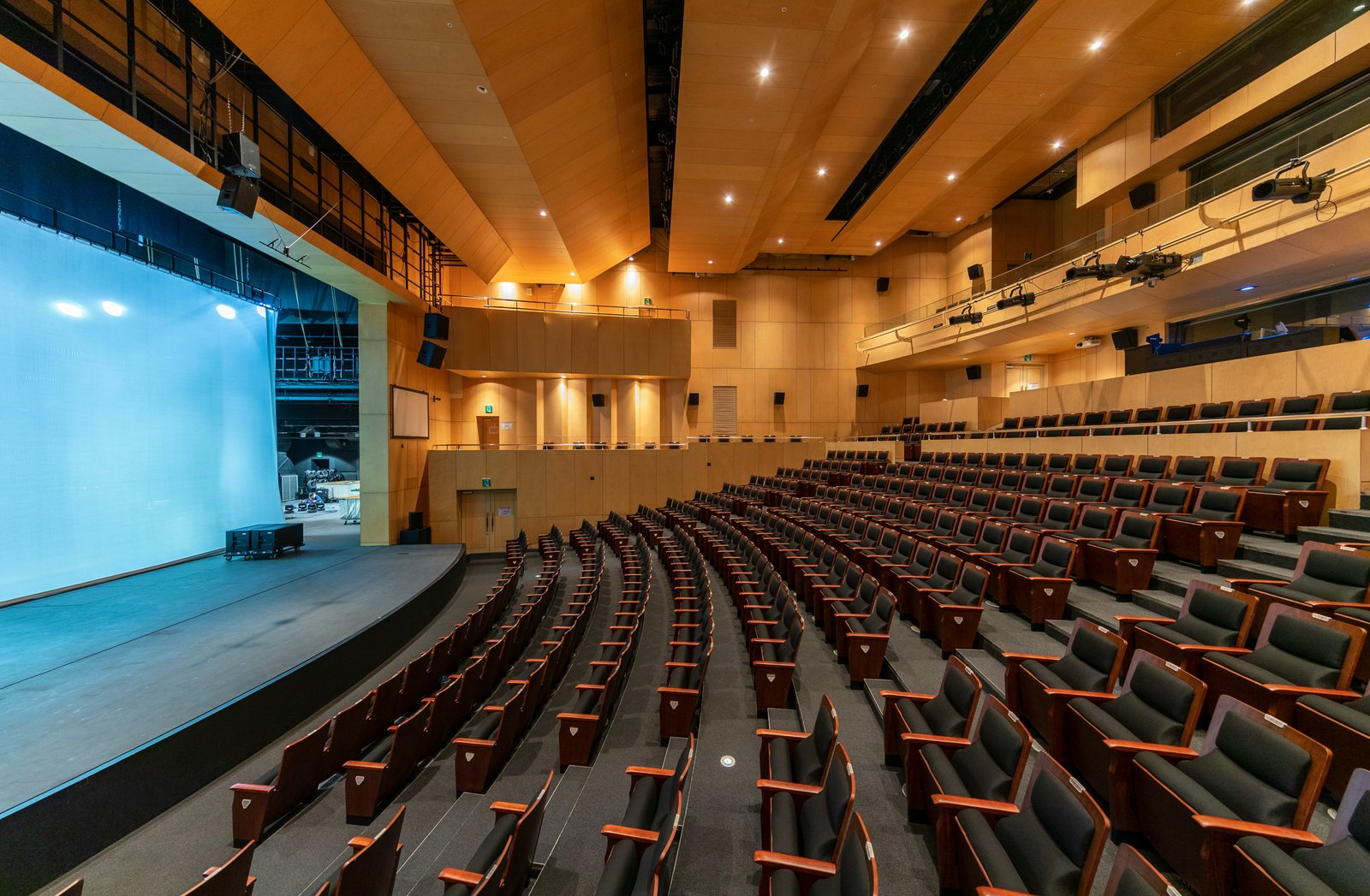 A wide interior view of a cinema theater showing rows of burgundy seats facing a lit projection screen and stage, with warm ambient lighting and modern architectural elements including recessed ceiling fixtures and mounted displays.