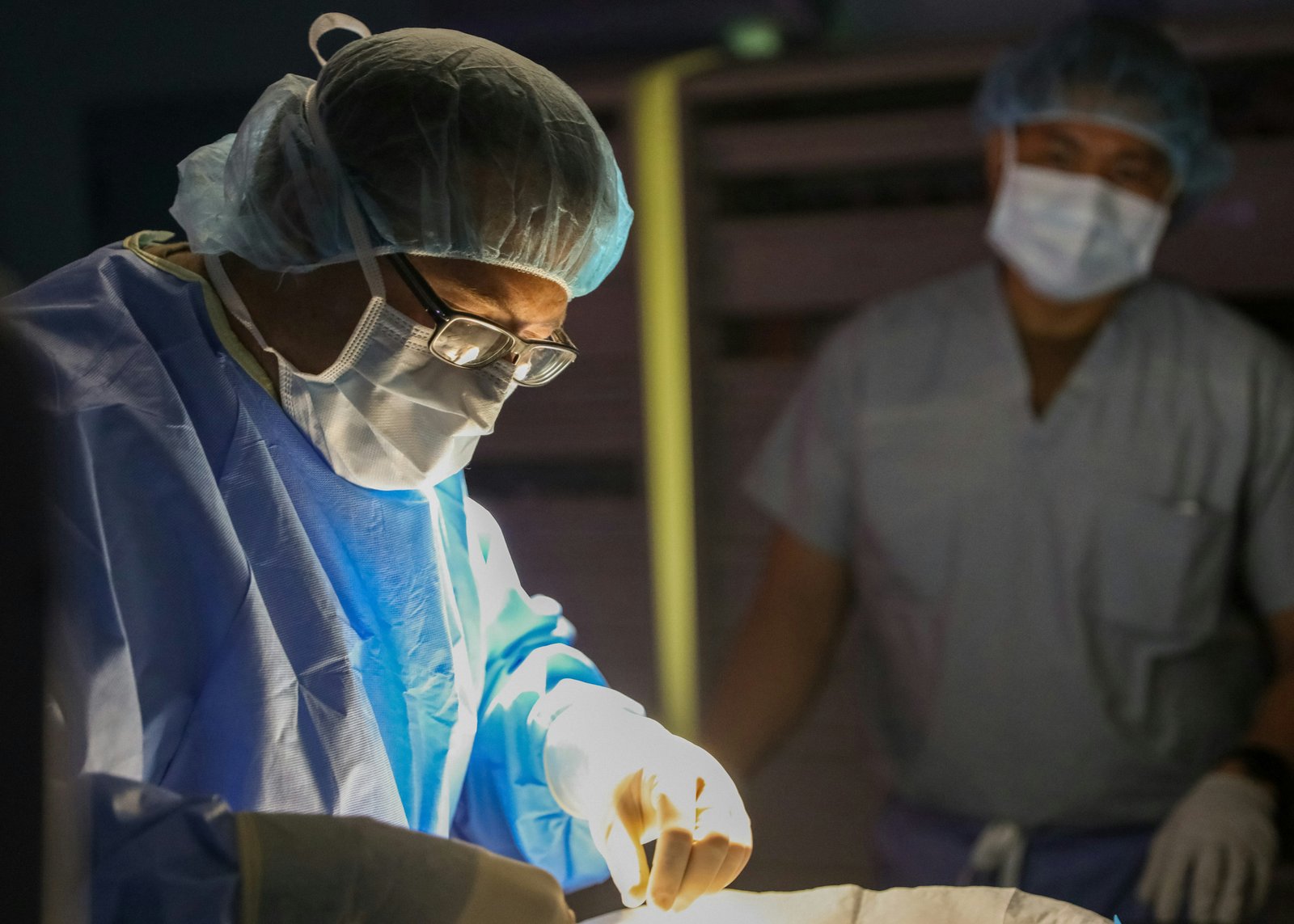 A surgeon wearing a head light, surgical cap, mask, and glasses performs a procedure on a sterile field while a second medical professional in protective gear observes in the background in an operating room setting.