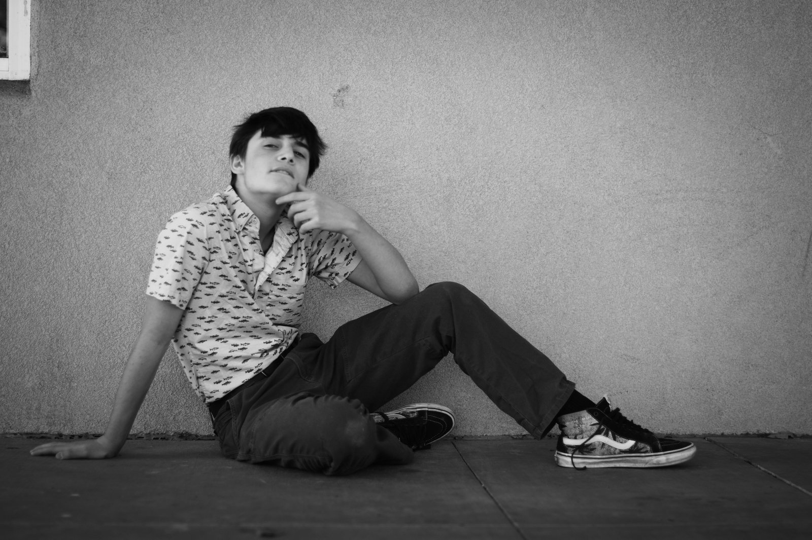 A black and white photograph of a young teenage person sitting alone on a floor against a plain wall, wearing a patterned short-sleeved shirt and dark pants with casual sneakers, with a contemplative and pensive facial expression while resting their head on their hand.
