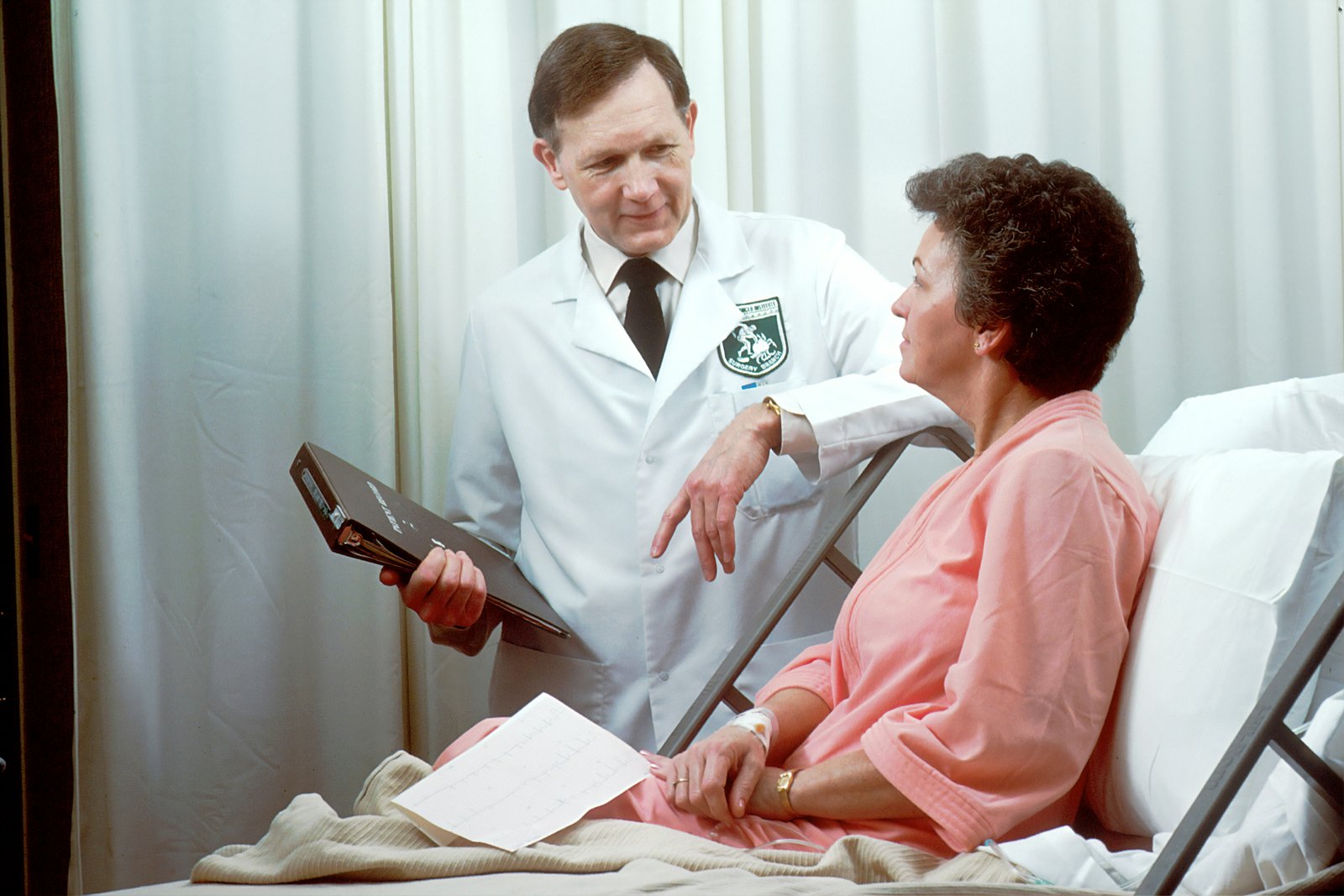 A male doctor in a white coat speaks with a female patient in a pink hospital gown while holding a tablet or clipboard, sitting together on a hospital bed in a clinical setting.