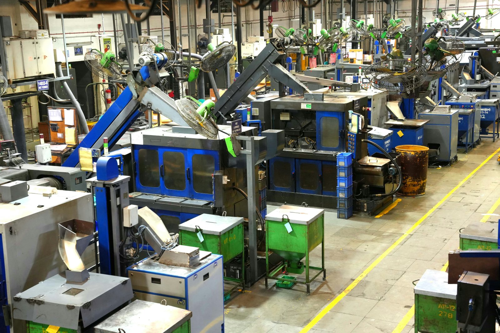 Wide landscape view of an industrial manufacturing facility floor with blue and black heavy machinery, green hoppers, and equipment arranged throughout a large warehouse space with yellow safety lines painted on the concrete floor.