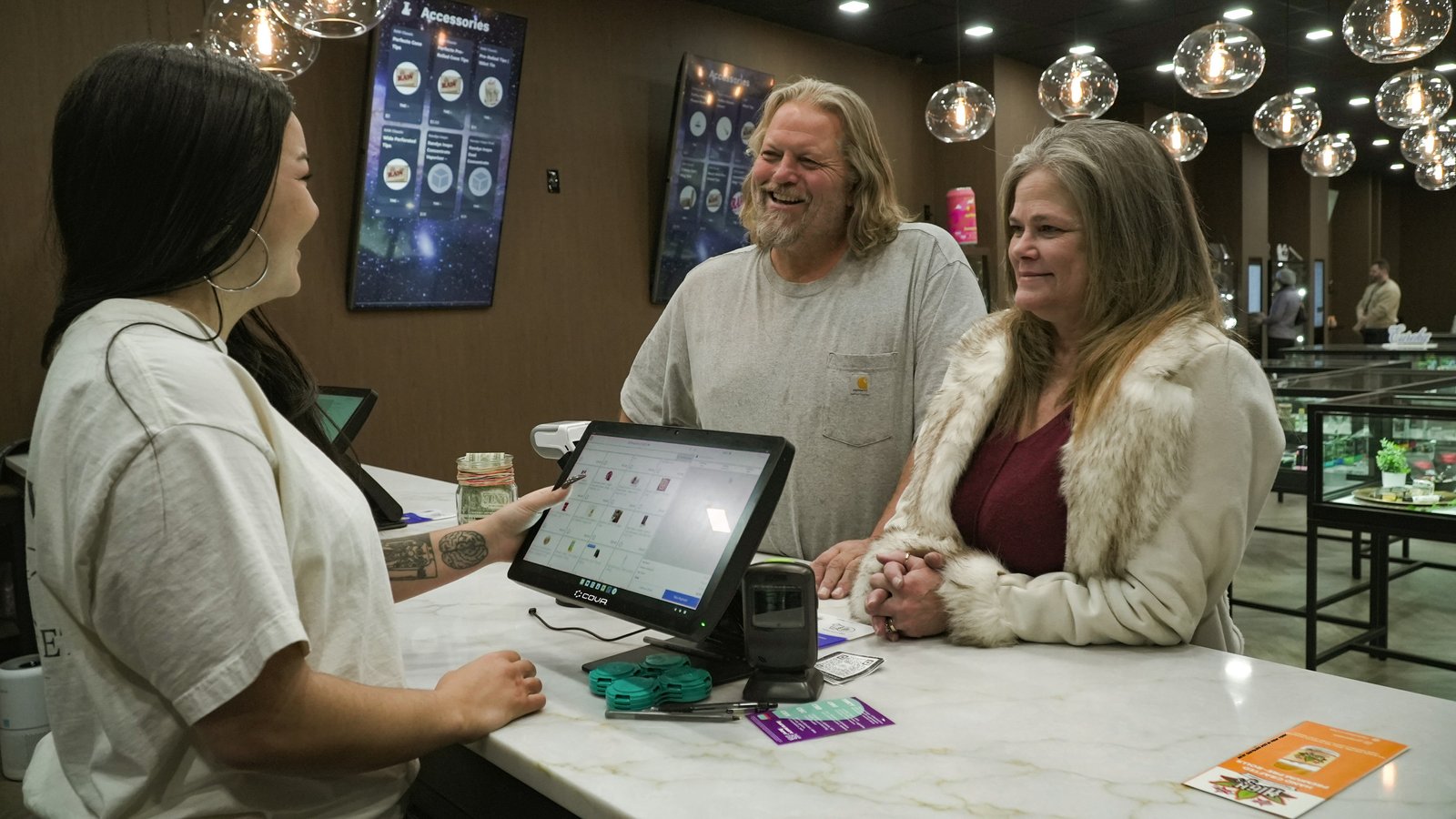 A jewelry store employee assists two customers at a counter with a tablet display while pendant lights hang overhead in the showroom.