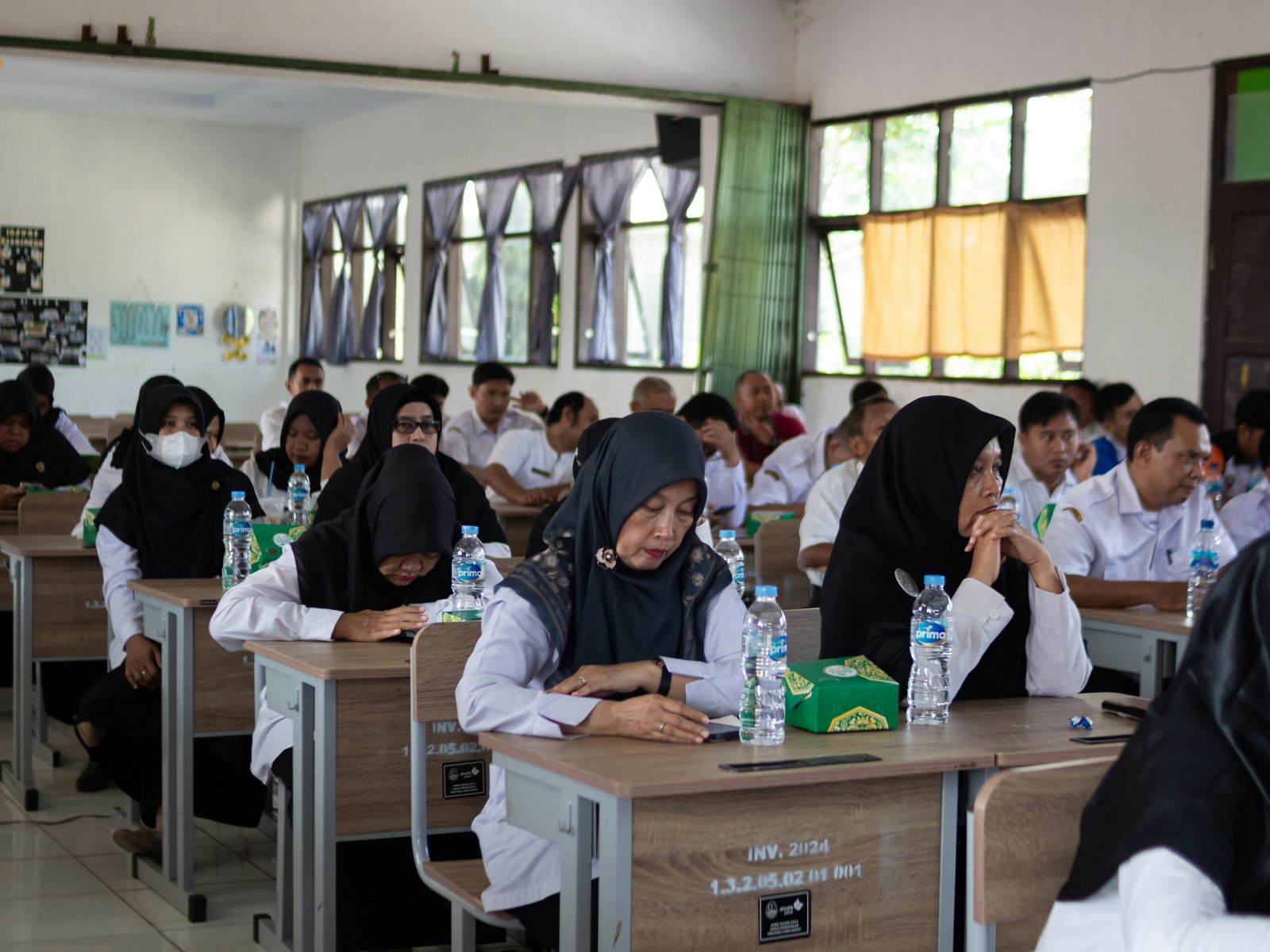 Students in school uniforms sitting at individual desks in a classroom working on assignments, with water bottles on their desks and natural light coming through windows in the background.