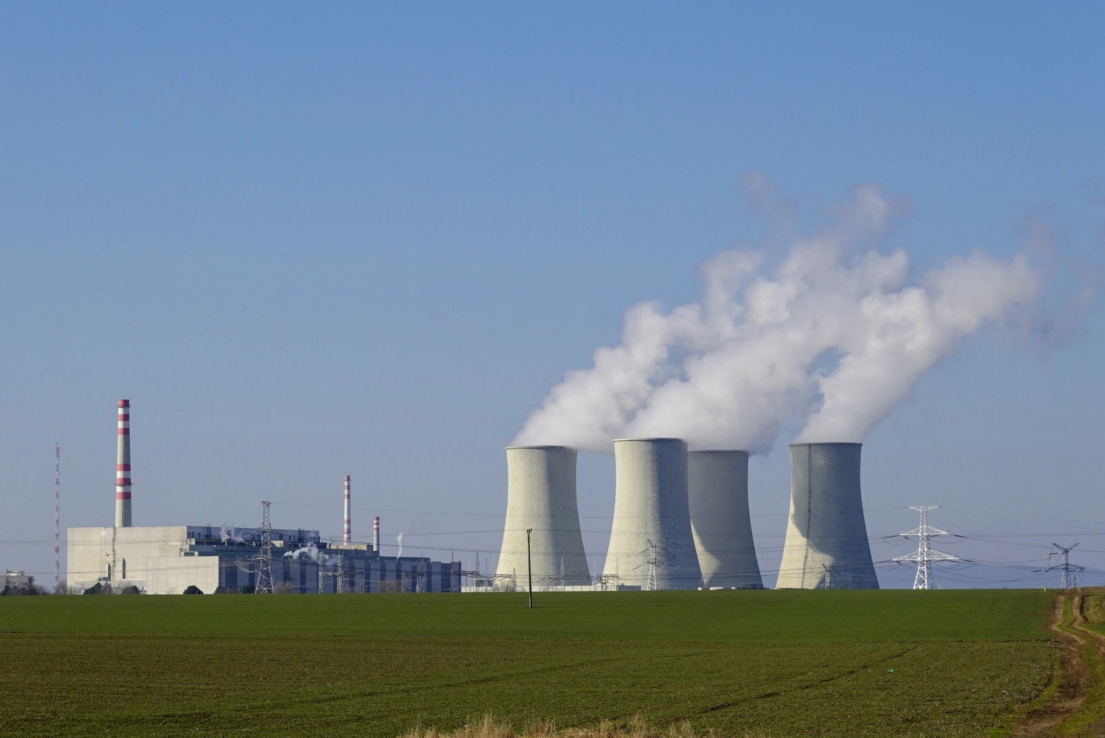 A coal-fired power plant with four large cooling towers emitting white steam plumes stands in a flat agricultural landscape with green fields, accompanied by industrial structures and transmission lines under a partly cloudy sky.