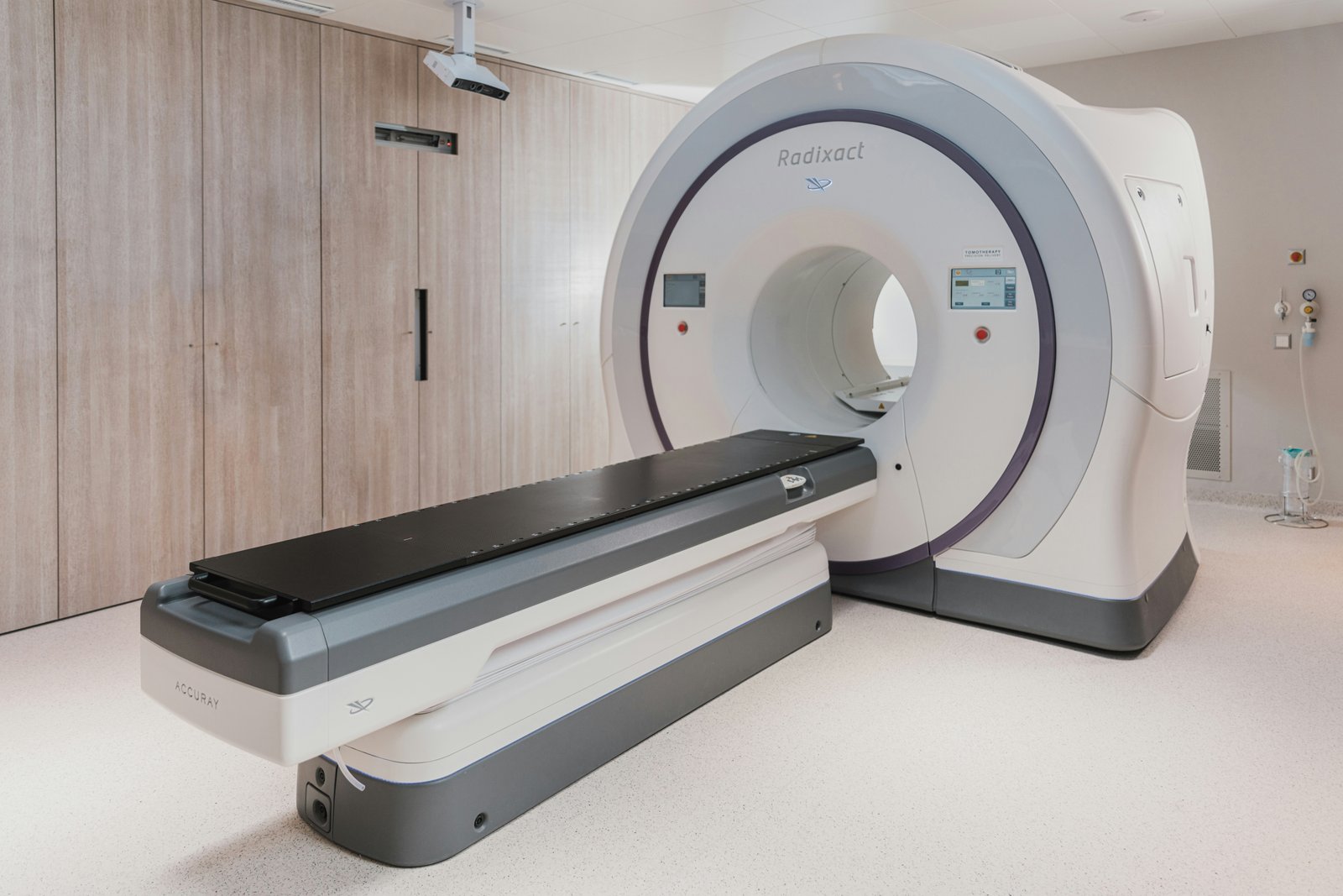 A white and gray CT scanner with an open gantry and patient table in a bright, clean hospital radiology department room with wooden cabinetry visible in the background.