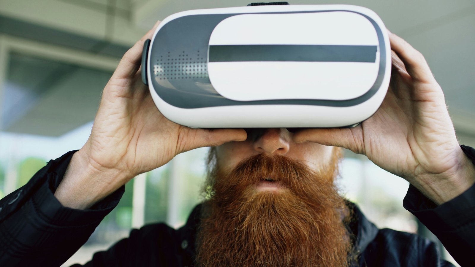 A bearded man with brown hair, wearing a black jacket, holds a white virtual reality headset up to his face with both hands in an indoor setting with a blurred background.