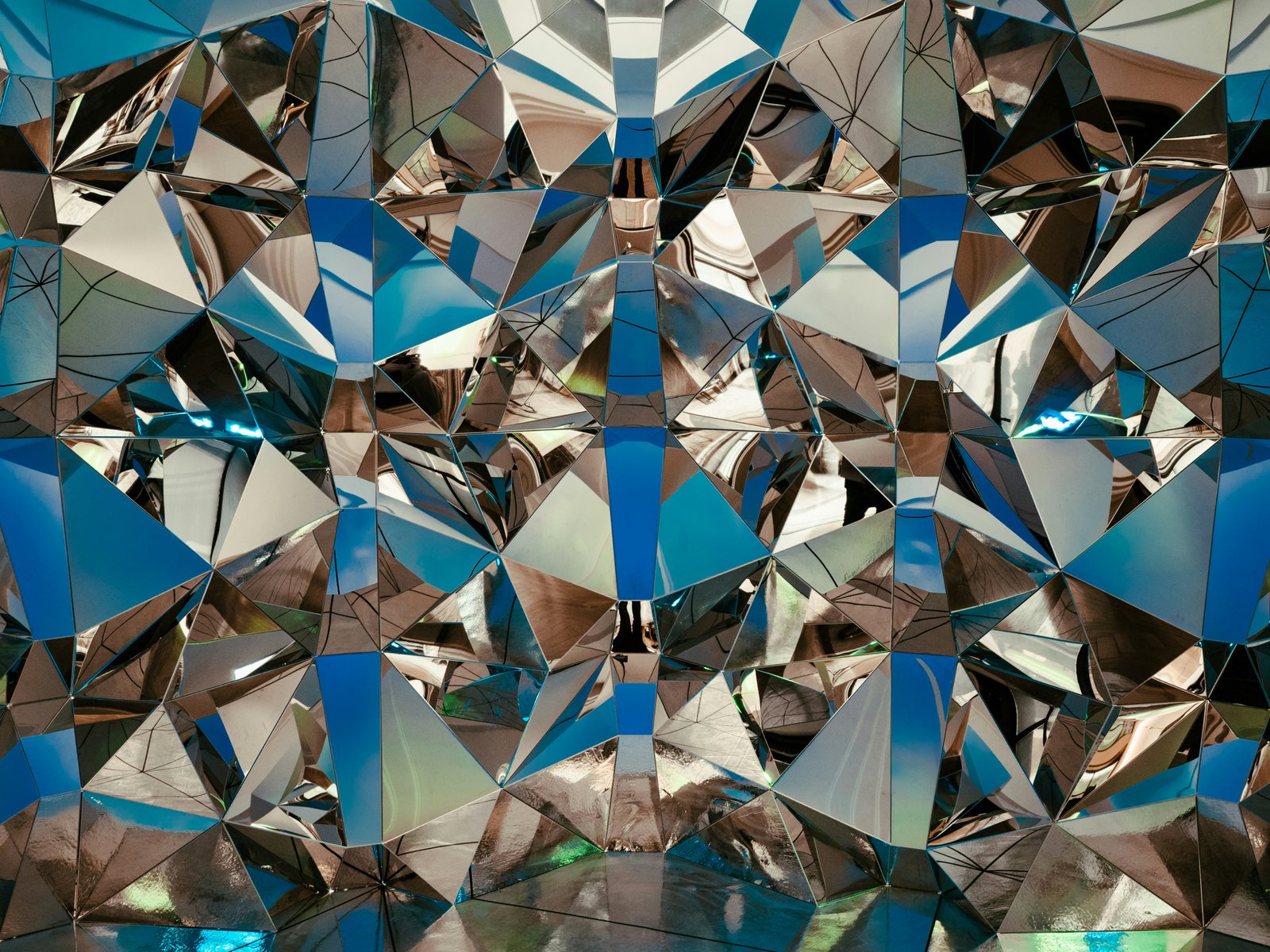 A complex geometric pattern of angular, reflective facets in blue, silver, brown, and white tones creates a fragmented, kaleidoscopic mosaic effect.