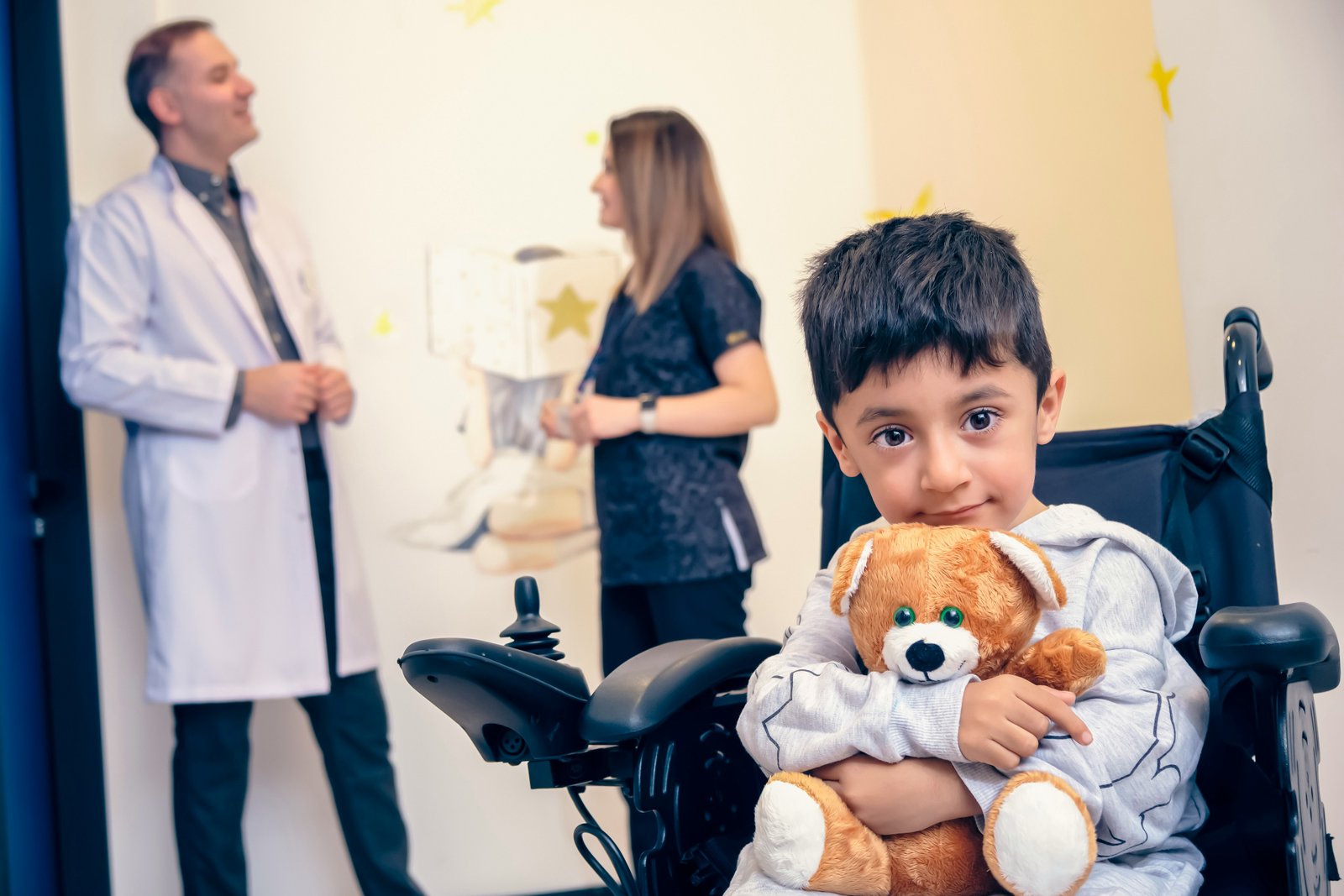 A young boy in a wheelchair holding a teddy bear sits in the foreground of a clinic examination room, while a male doctor in a white coat and a female healthcare worker in dark clothing confer in the background against a pale yellow wall.