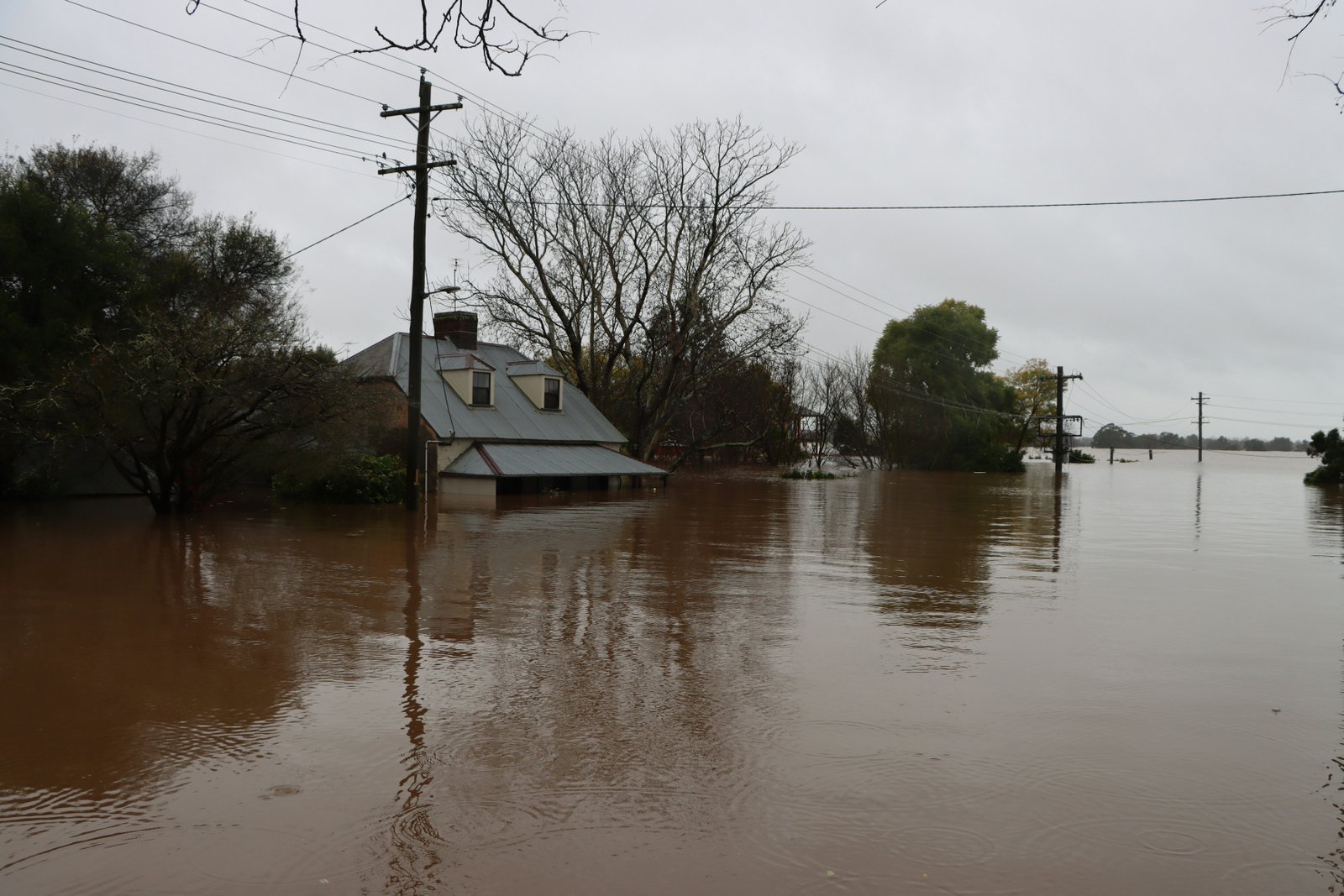 A residential area flooded with brown water, showing a house and utility poles partially submerged in an expansive flood plain landscape.