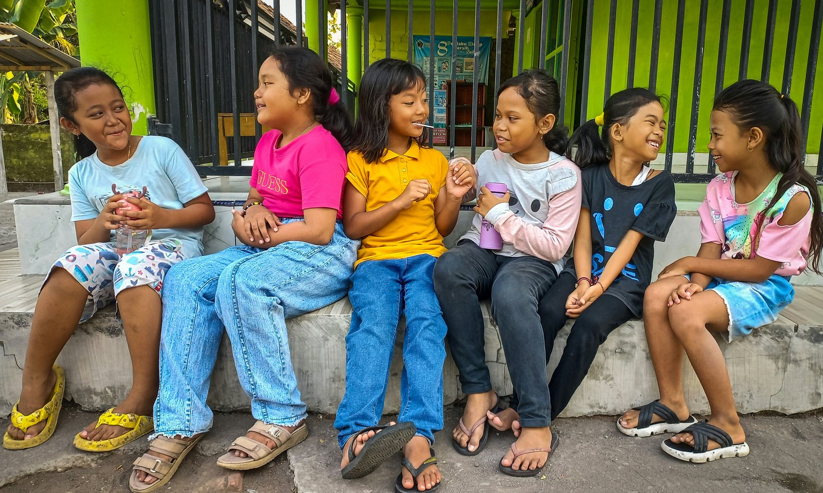 Six children of various ages sit together on a concrete step outside a simple green building, wearing casual colorful clothing, appearing to engage in a group conversation or gathering in a community setting.