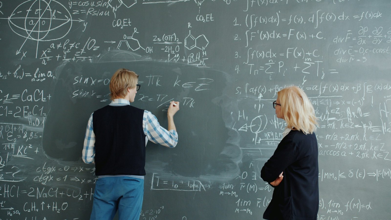 Two people stand at a large chalkboard covered in mathematical equations and formulas, with one person in a black vest and jeans writing on the board while another person in a black blazer looks on.