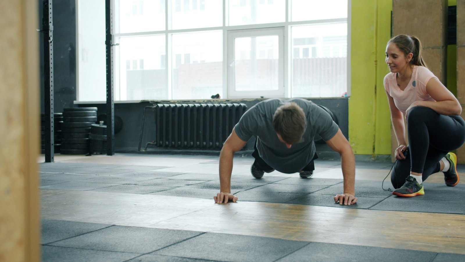 Two people performing plank exercises on mats in a bright, modern gymnasium with large windows, industrial flooring, and a radiator visible in the background.
