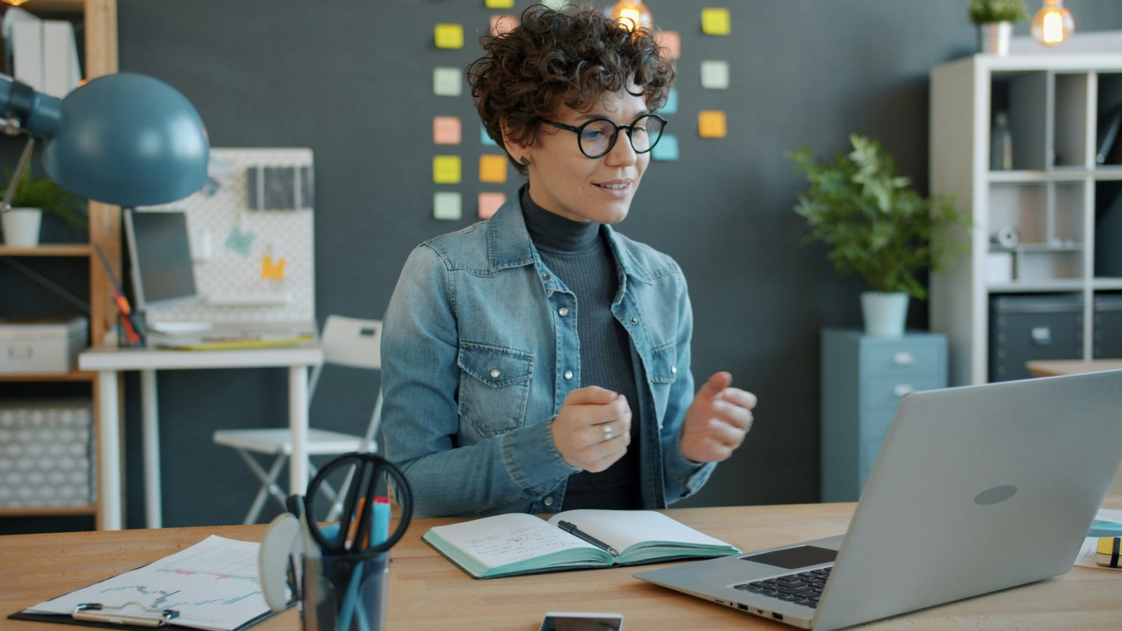 A person wearing glasses and a denim shirt sits at a wooden desk in a modern office, gesturing while looking at a laptop screen, with office furniture and storage visible in the background.