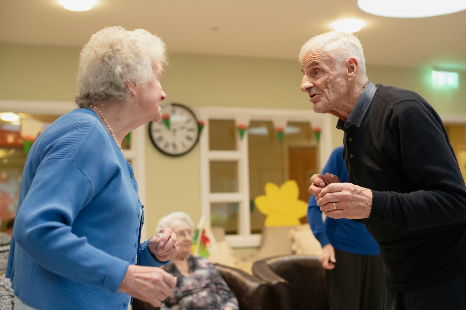 Two elderly people engaged in conversation in an institutional interior space with a wall clock, fluorescent lighting, and doorways visible in the background.