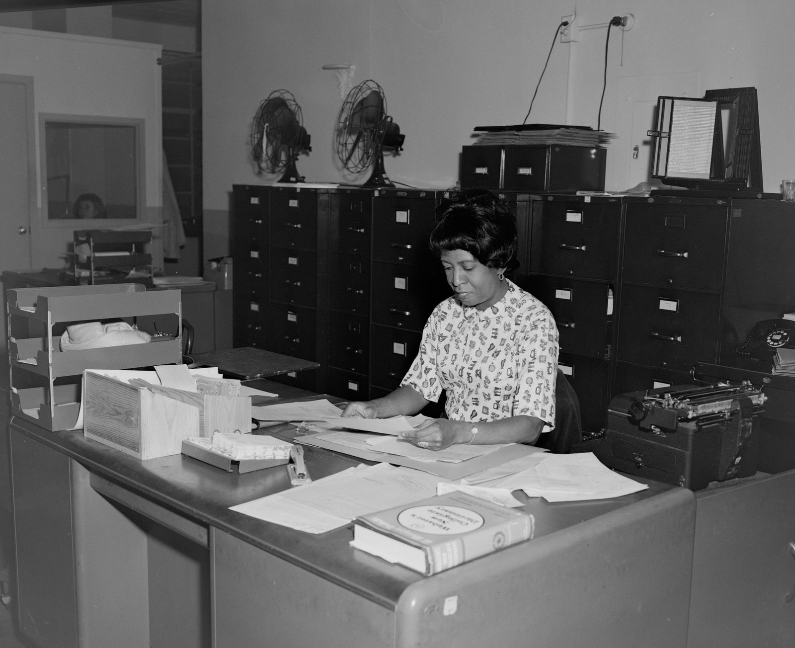 A black and white photograph of a woman sitting at a desk in what appears to be a government office, surrounded by filing cabinets and paperwork, engaged in administrative work.