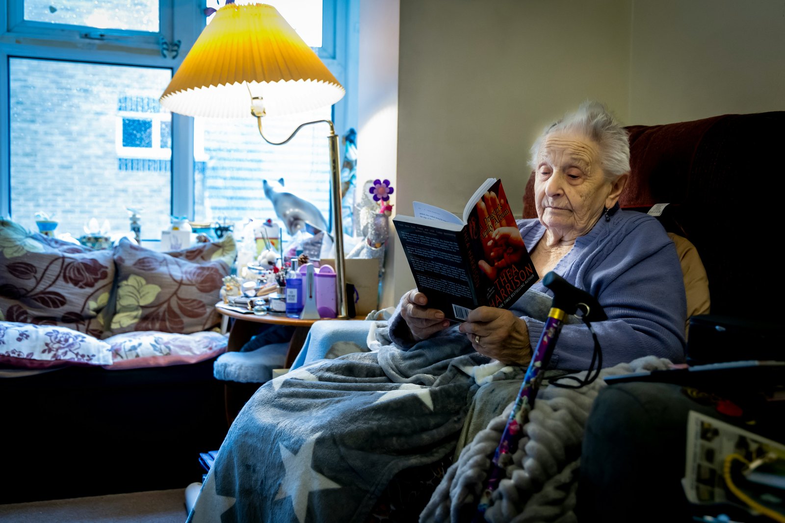 An elderly person sits in a comfortable chair in a home or care room setting, holding a tablet device, with a yellow desk lamp and personal items visible in the background, suggesting a quiet, intimate care environment.