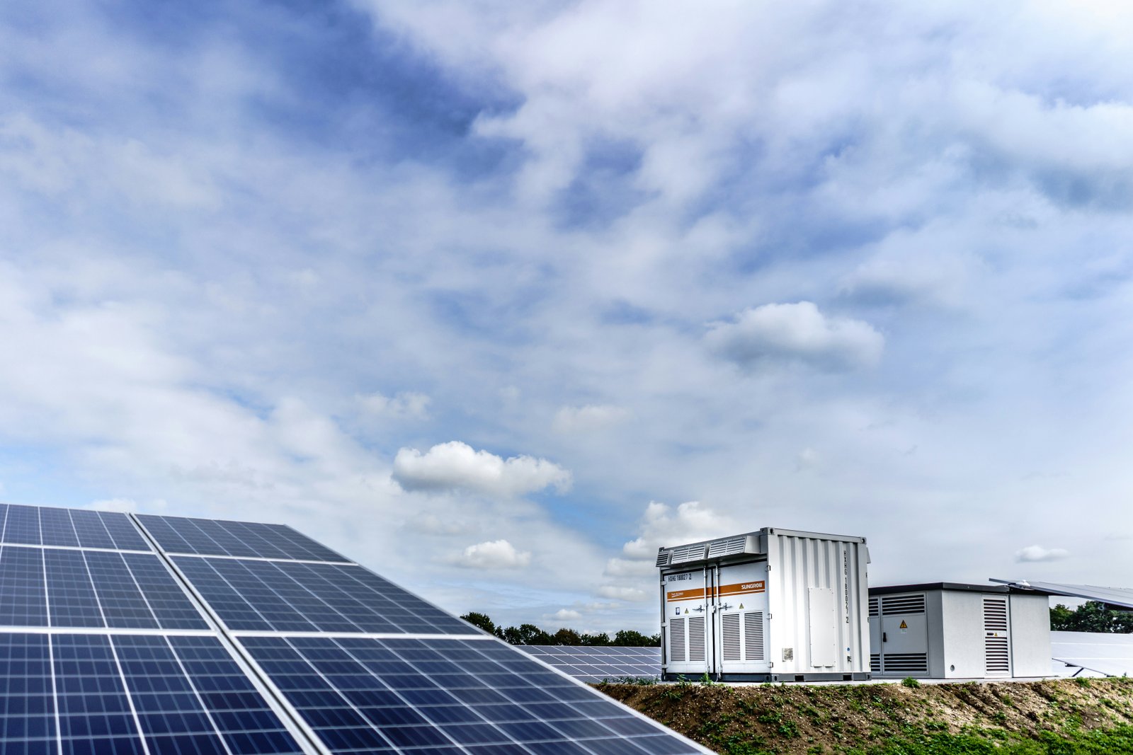 A landscape photograph showing a large array of solar panels in the foreground with a modern white industrial or manufacturing facility building visible in the background under a partly cloudy blue sky.