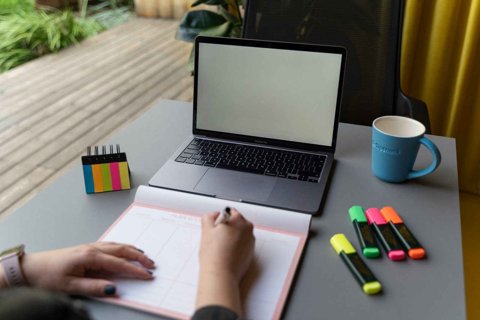 A person's hands write in an open notebook at a desk with a laptop, colorful highlighters, sticky notes, and a blue mug.