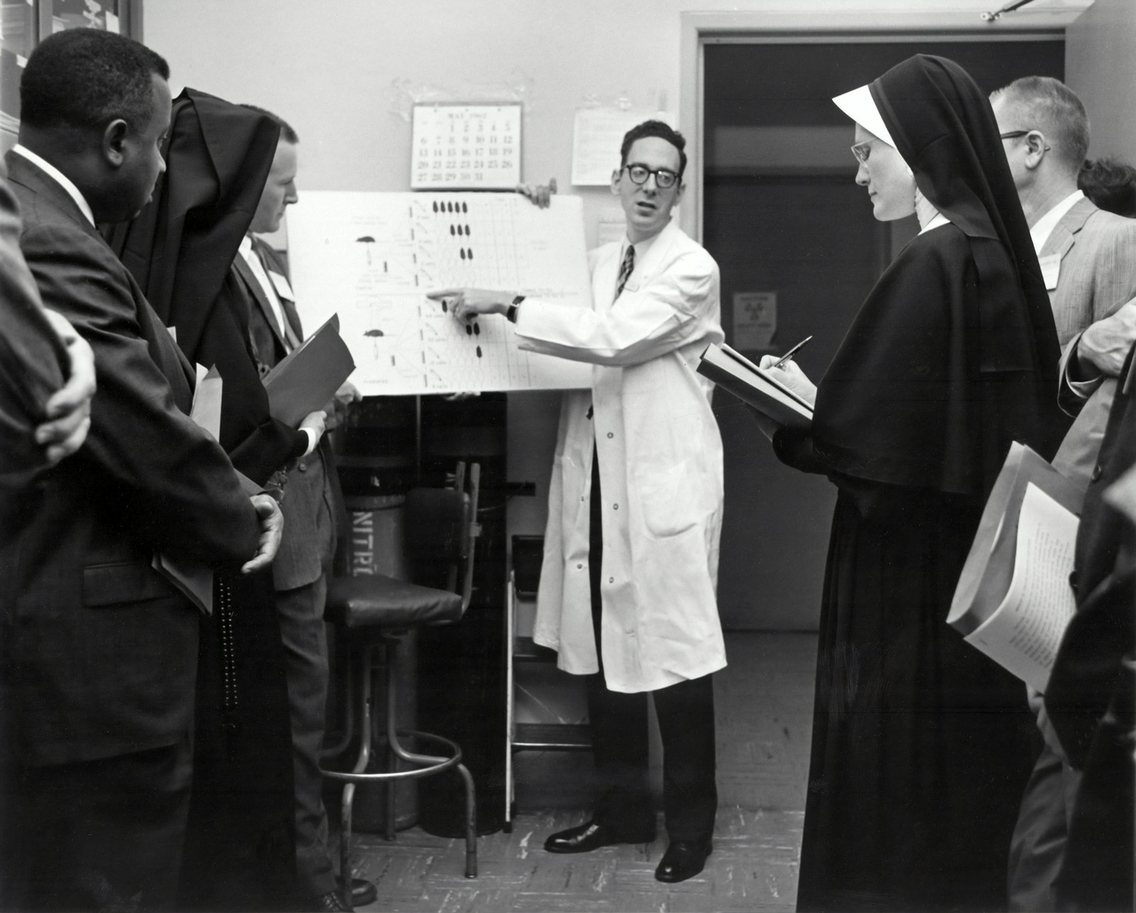 A black and white photograph of healthcare professionals in a clinical setting gathered around a woman in a white coat who is pointing to or presenting information on a wall-mounted chart or document, with several men in suits observing and engaged in discussion.