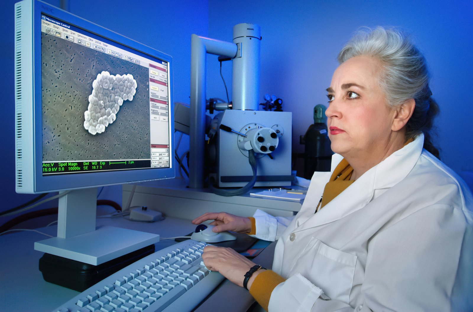 A woman in a white lab coat sits at a computer workstation in a laboratory, viewing what appears to be microscopic or medical imaging data on a monitor, with laboratory equipment including a microscope visible in the background against blue lighting.