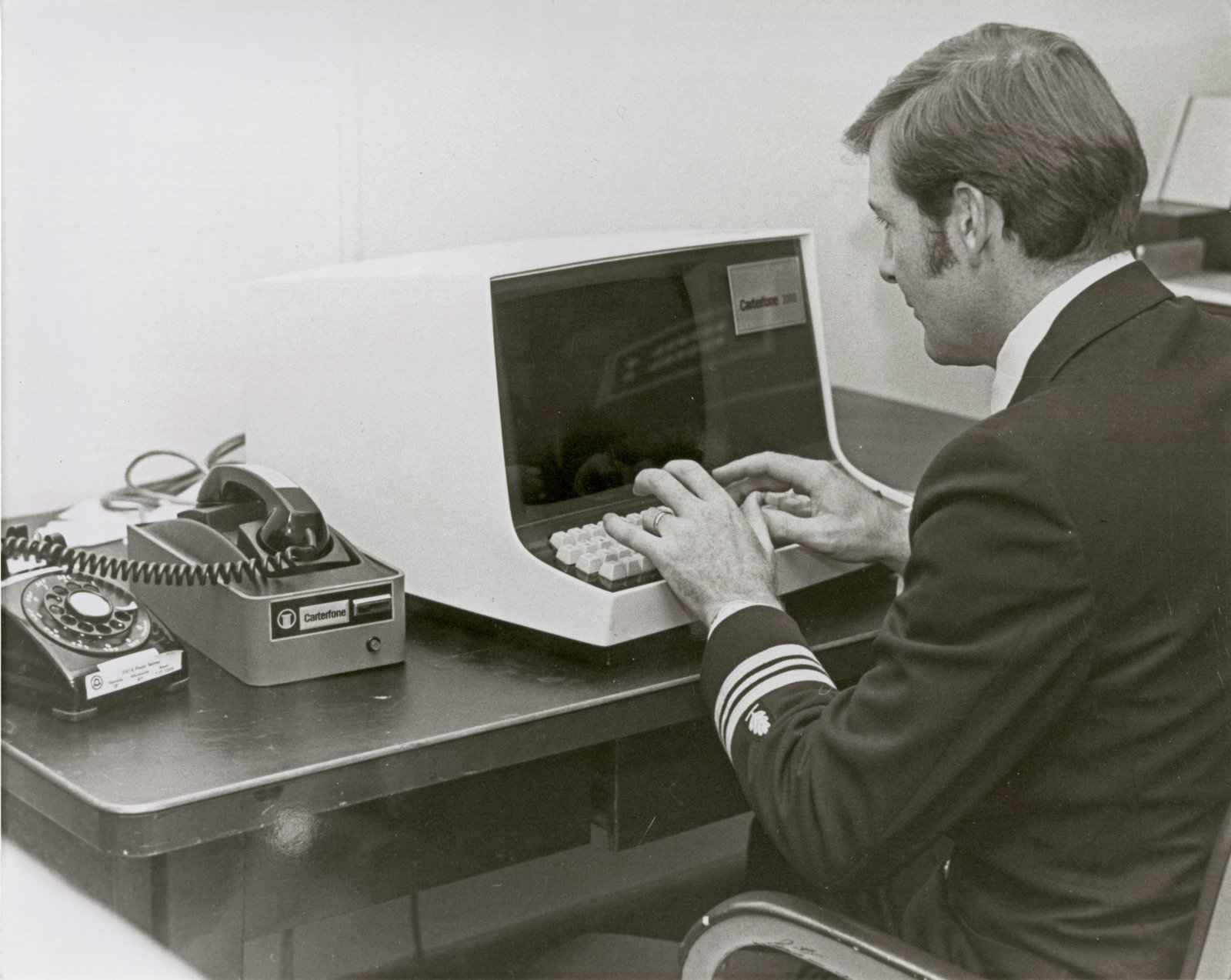 A man in a dark business suit sits at a desk reviewing information on a large computer monitor, with office equipment visible to his left in a clinical or professional setting.