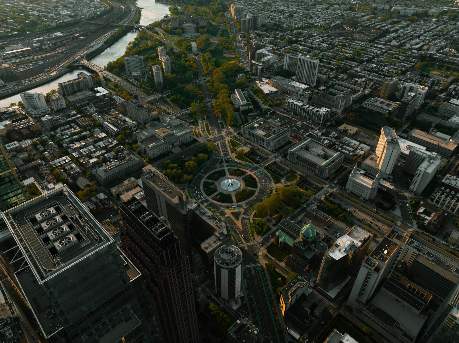 Aerial photograph of a dense urban cityscape with tall buildings, streets, and a prominent circular plaza or roundabout in the center, surrounded by arid terrain and visible urban sprawl extending to the horizon.