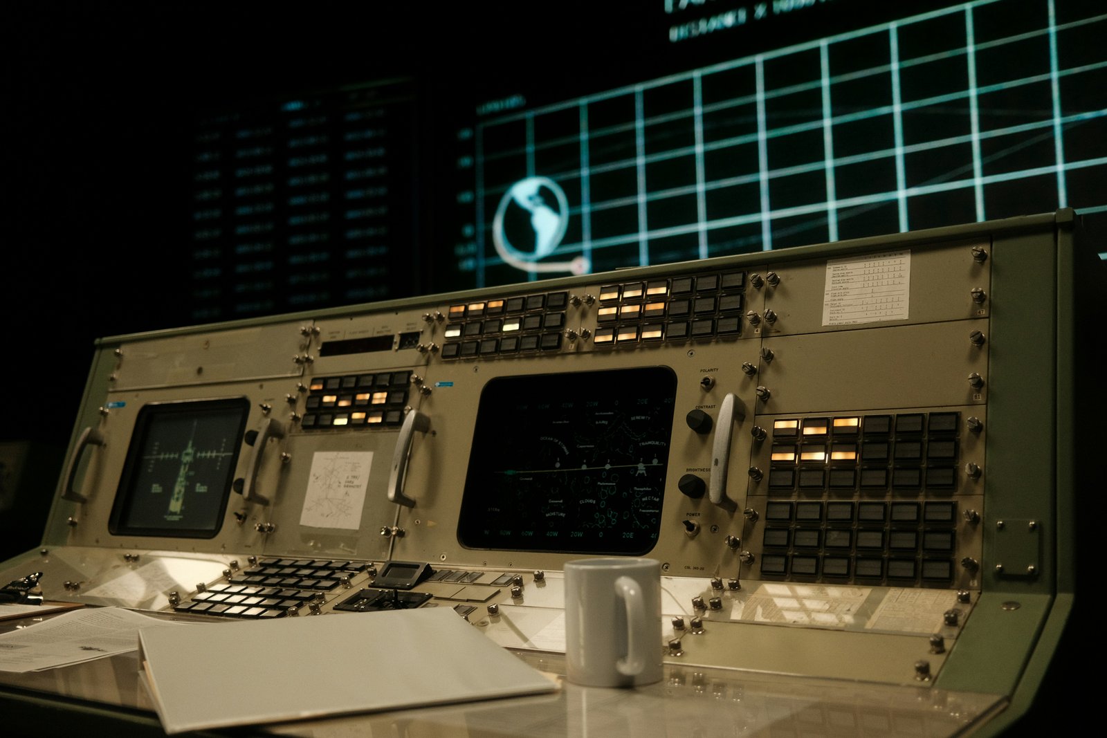 A vintage spacecraft or aircraft control panel with illuminated instrumentation, gauges, switches, and display screens arranged across a beige console, photographed against a dark background with a gridded screen visible above.