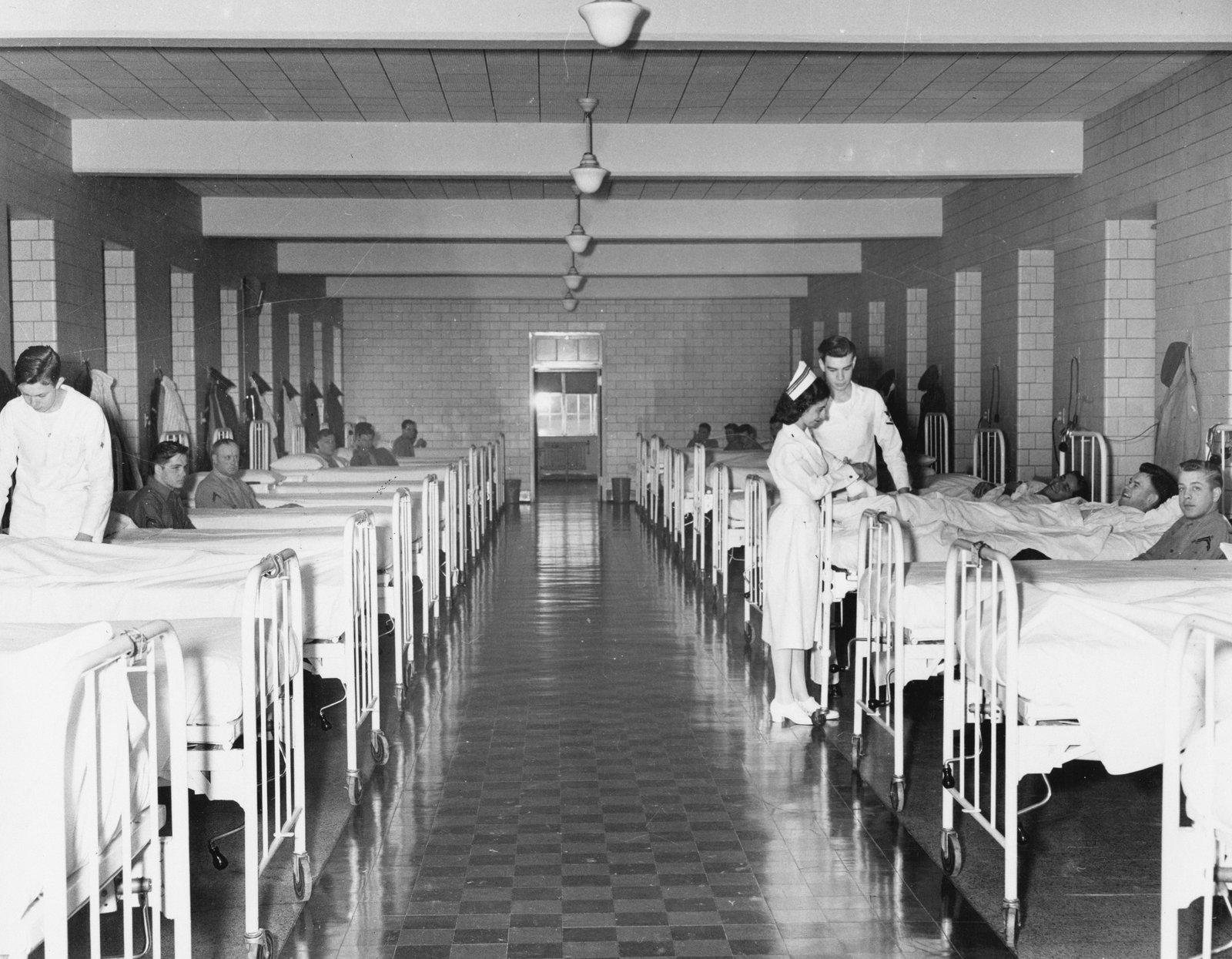 A black and white photograph showing a long hospital ward with two rows of metal-frame beds with white linens arranged along both walls, nurses in white uniforms attending to patients in their beds, and a checkered floor pattern extending down the center aisle with overhead lighting.