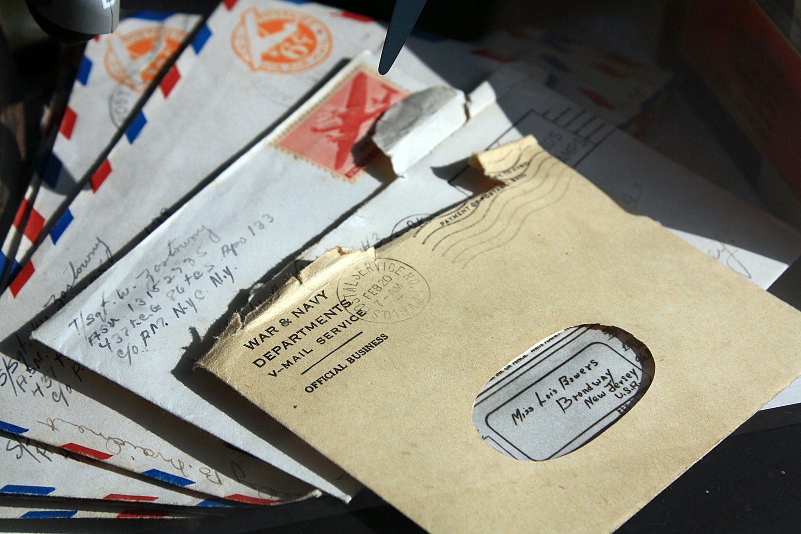 A collection of vintage letters and envelopes arranged on a dark surface, including a tan War & Navy Departments V-Mail Service envelope addressed to Miss Lois Bowers in Broadway, New Jersey, and other airmail correspondence with visible postmarks and handwritten addresses.