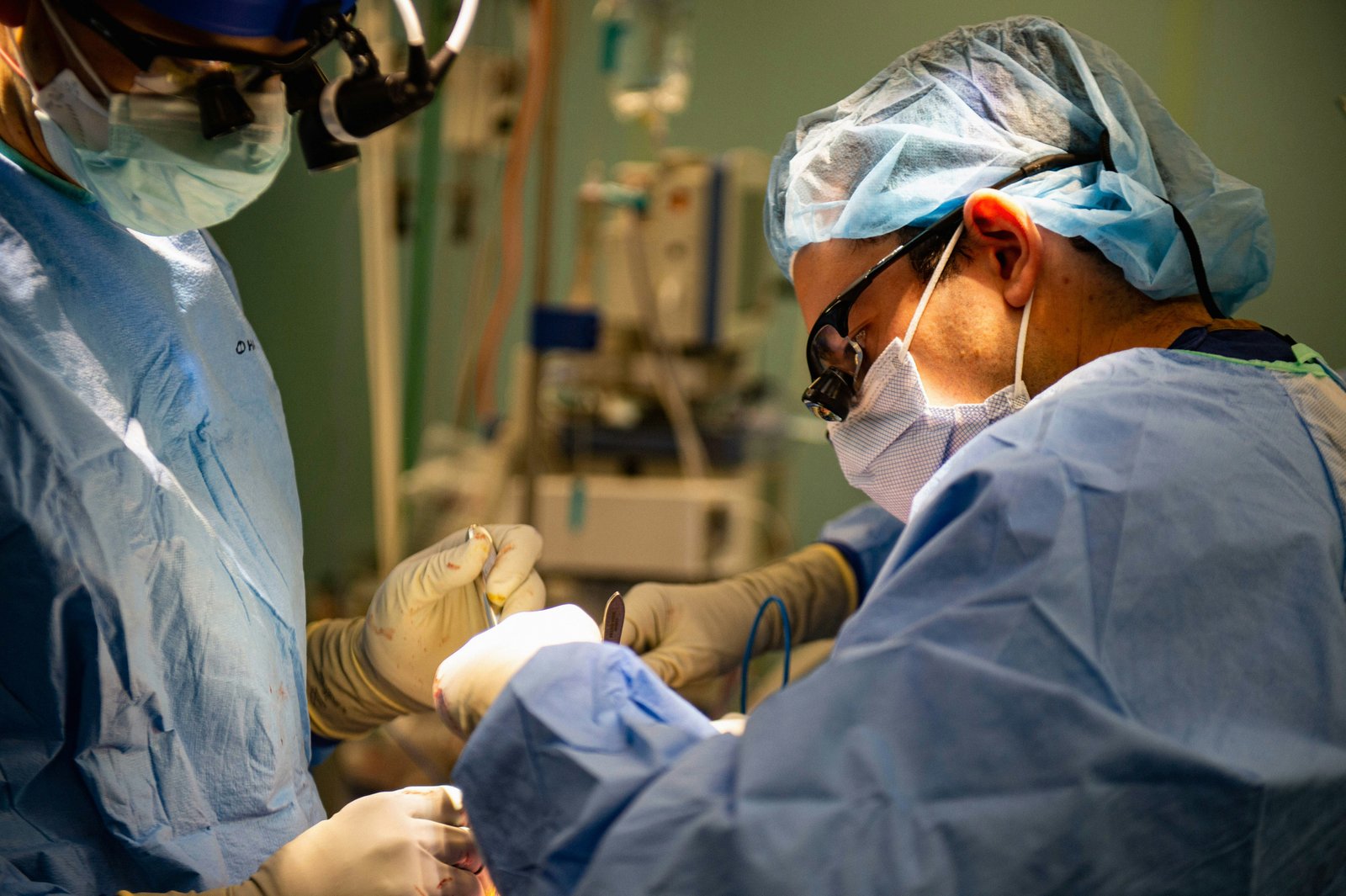 Two surgeons in protective eyewear and surgical attire working together in an operating room during a surgical procedure, with medical equipment and monitors visible in the background.