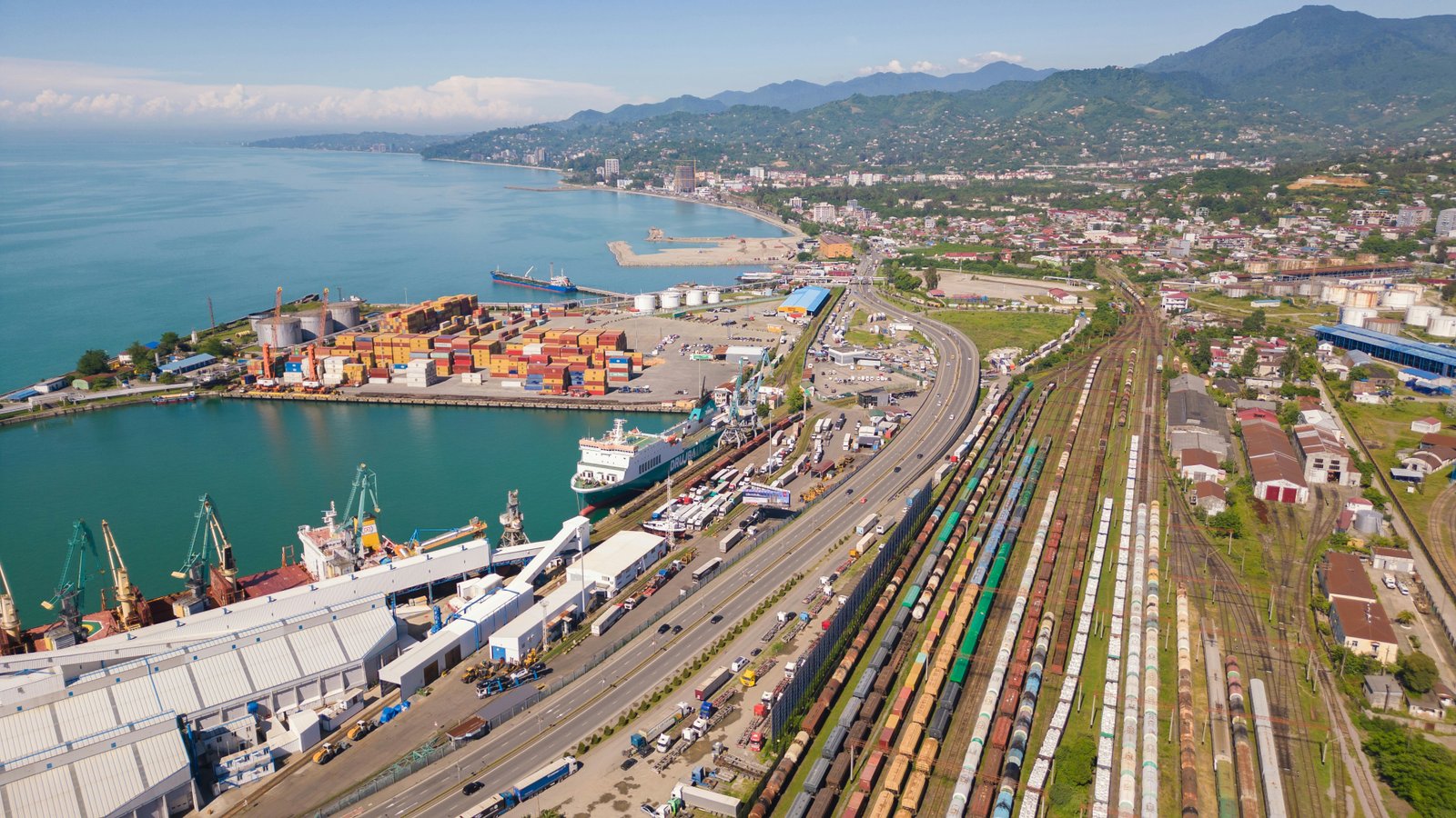 Aerial view of a modern cargo port terminal with container handling cranes, stacked shipping containers, docked vessels, railway tracks, and surrounding coastal residential areas with mountains in the distance.