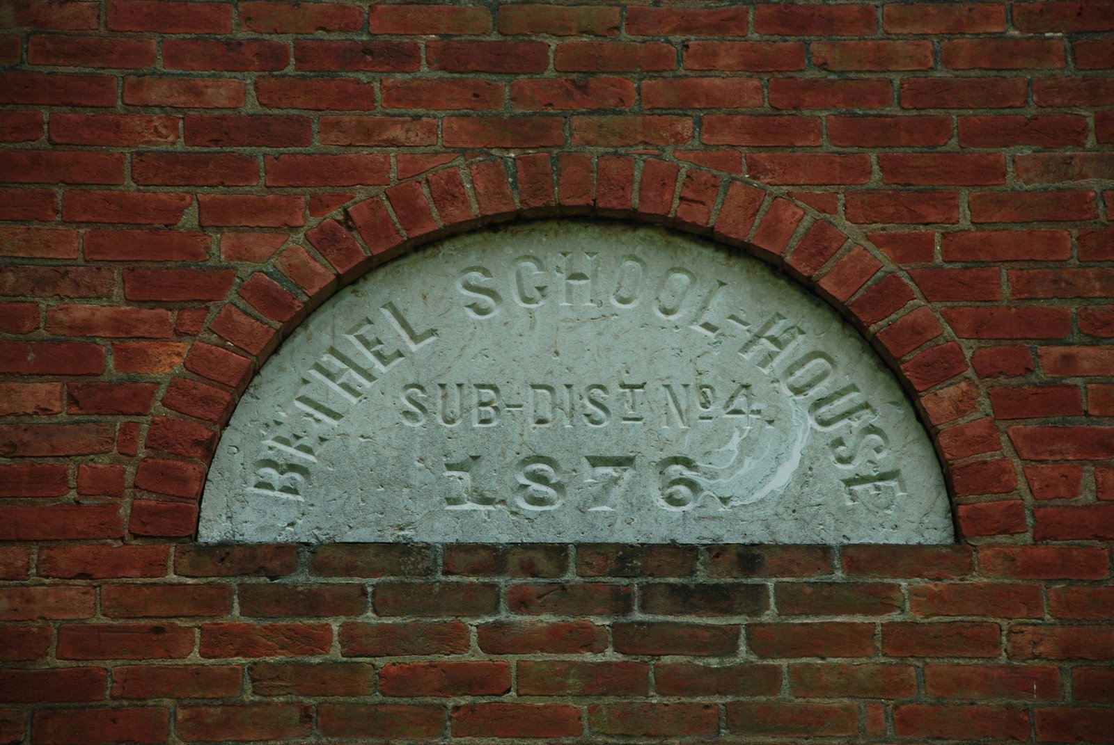 A semicircular stone plaque mounted on a red brick wall displaying text that reads "DANIEL SCHOOL-HOUSE SUB-DIST NO 4 1876", showing a historical educational institution nameplate.
