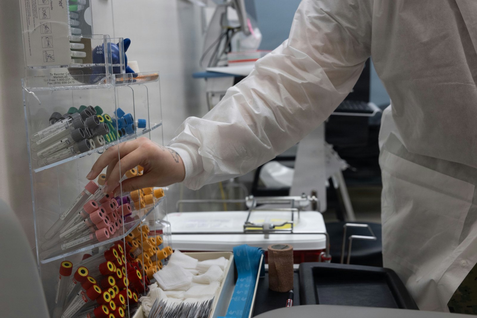 A healthcare worker in a white lab coat removes or organizes a red blood bag from a medical refrigeration unit in a laboratory setting, with laboratory equipment and storage visible in the background.