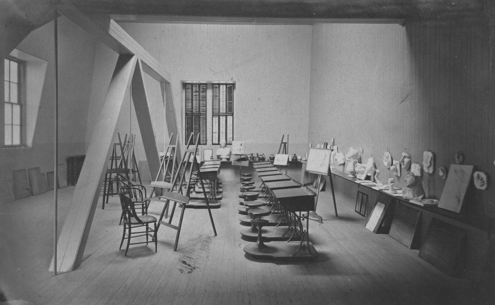A black and white photograph of a vintage mid-century classroom or studio space featuring long tables with papers and educational materials scattered across them, wooden chairs, easels, large windows with bars, and storage areas along the walls.