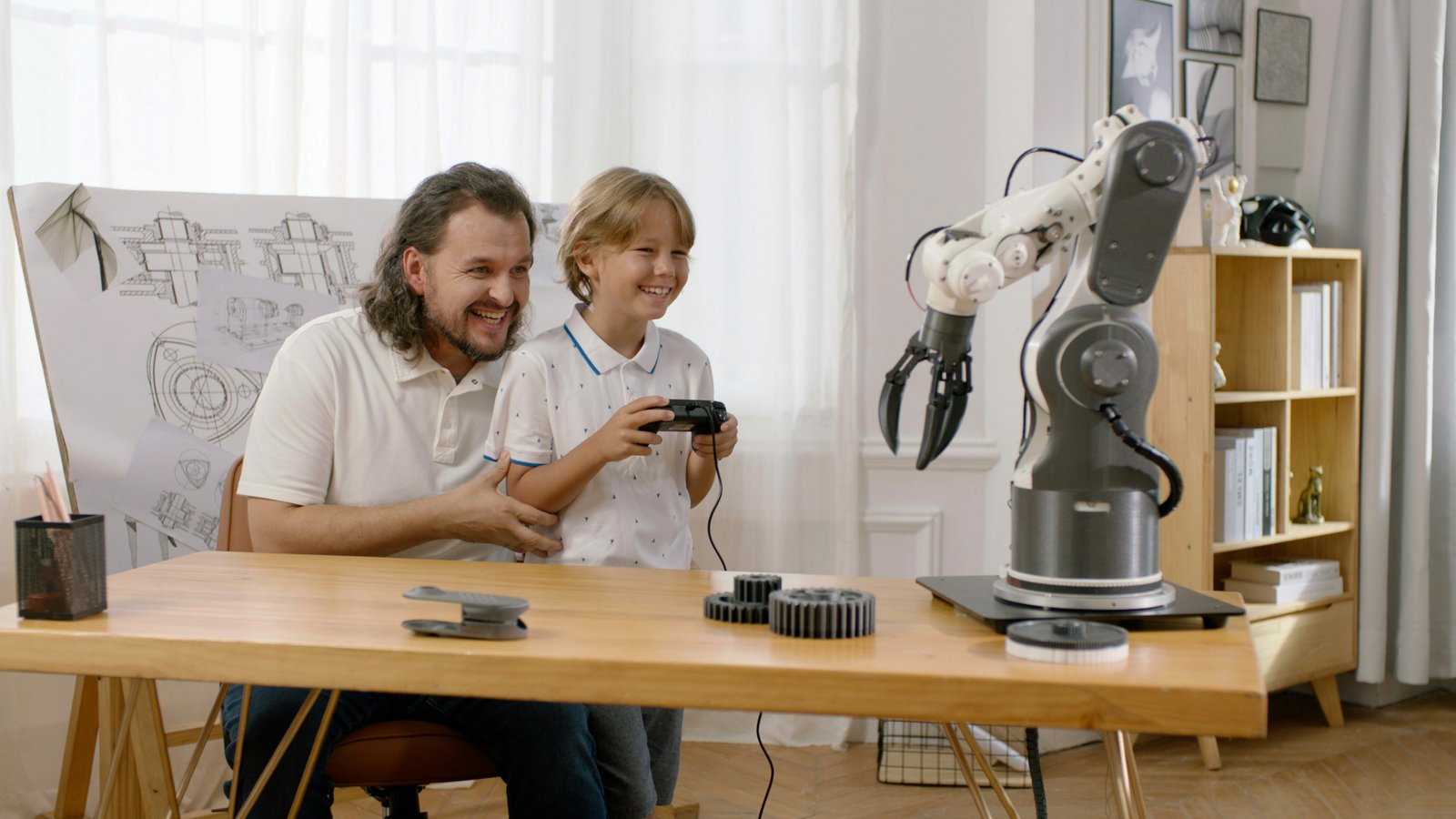 An adult man and young boy sit at a wooden classroom table working together with a robotic arm machine, with the boy holding a game controller while the man observes, in a bright classroom setting with shelving and a blueprint visible in the background.