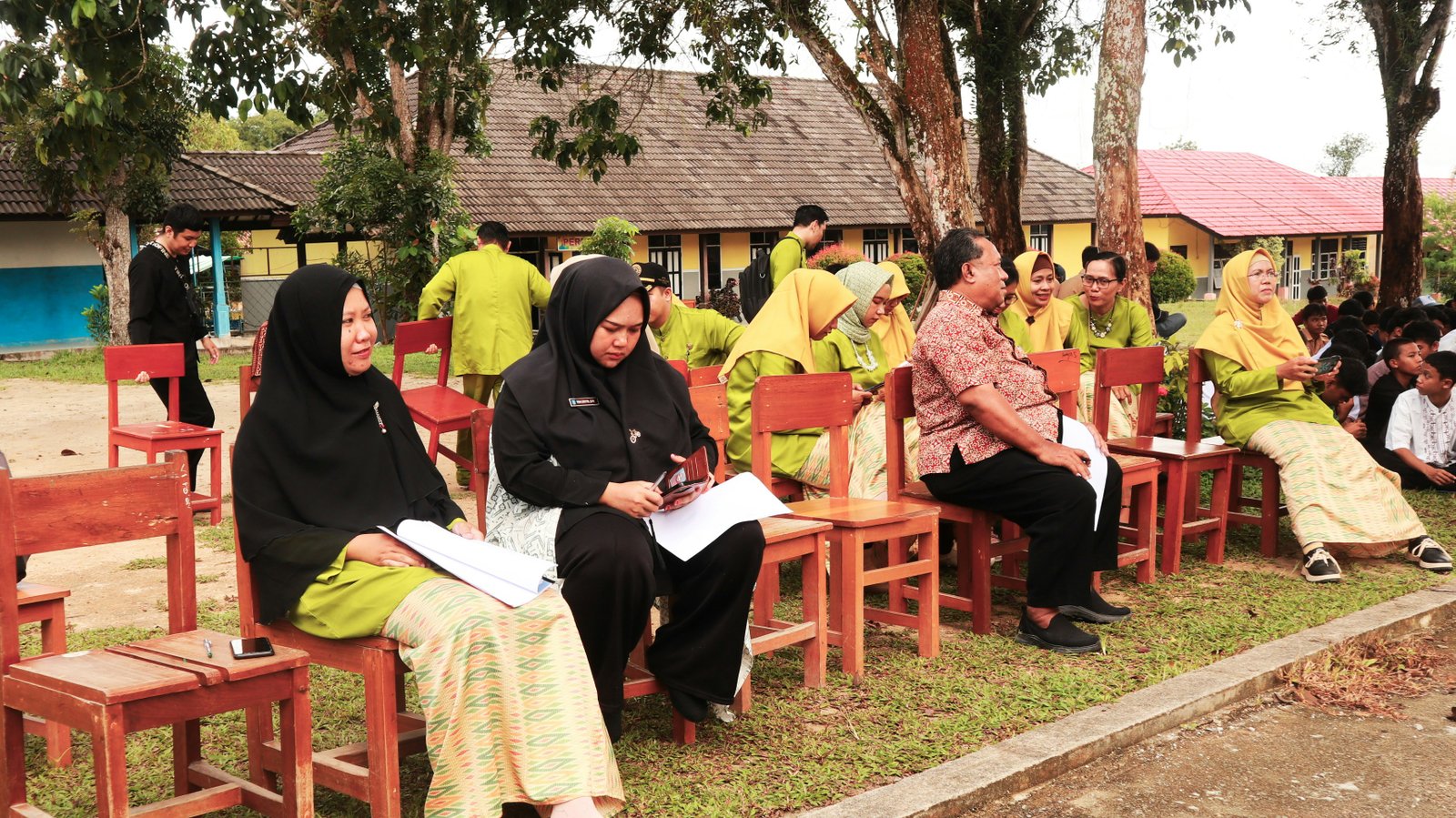 People sit on red wooden chairs and benches outdoors, with some wearing black hijabs and others in yellow and patterned clothing, with buildings and trees visible in the background.