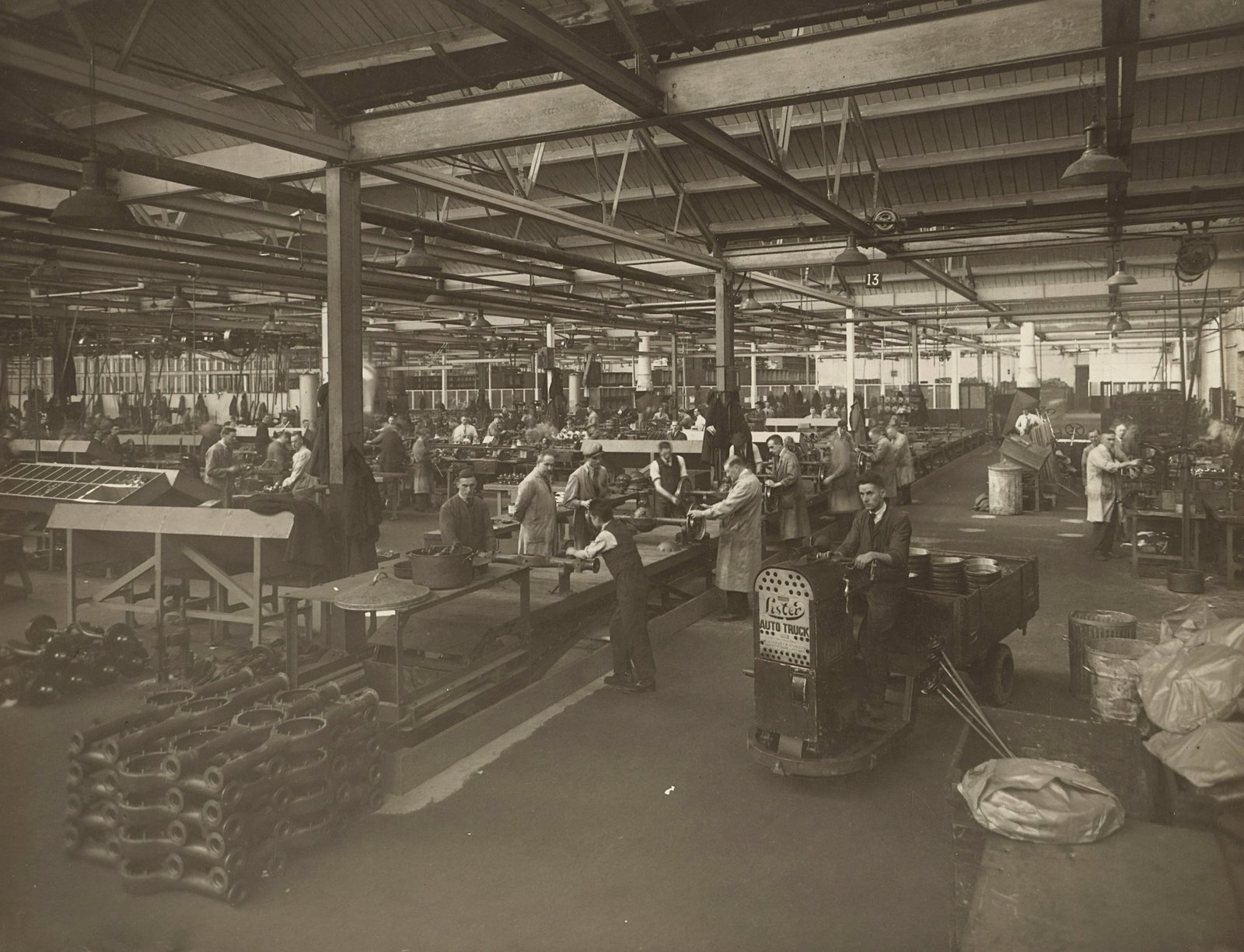 A black and white photograph of a large industrial factory floor with overhead steel beam structures, showing numerous workers scattered throughout the space among machinery and equipment, depicting historical wartime manufacturing work.