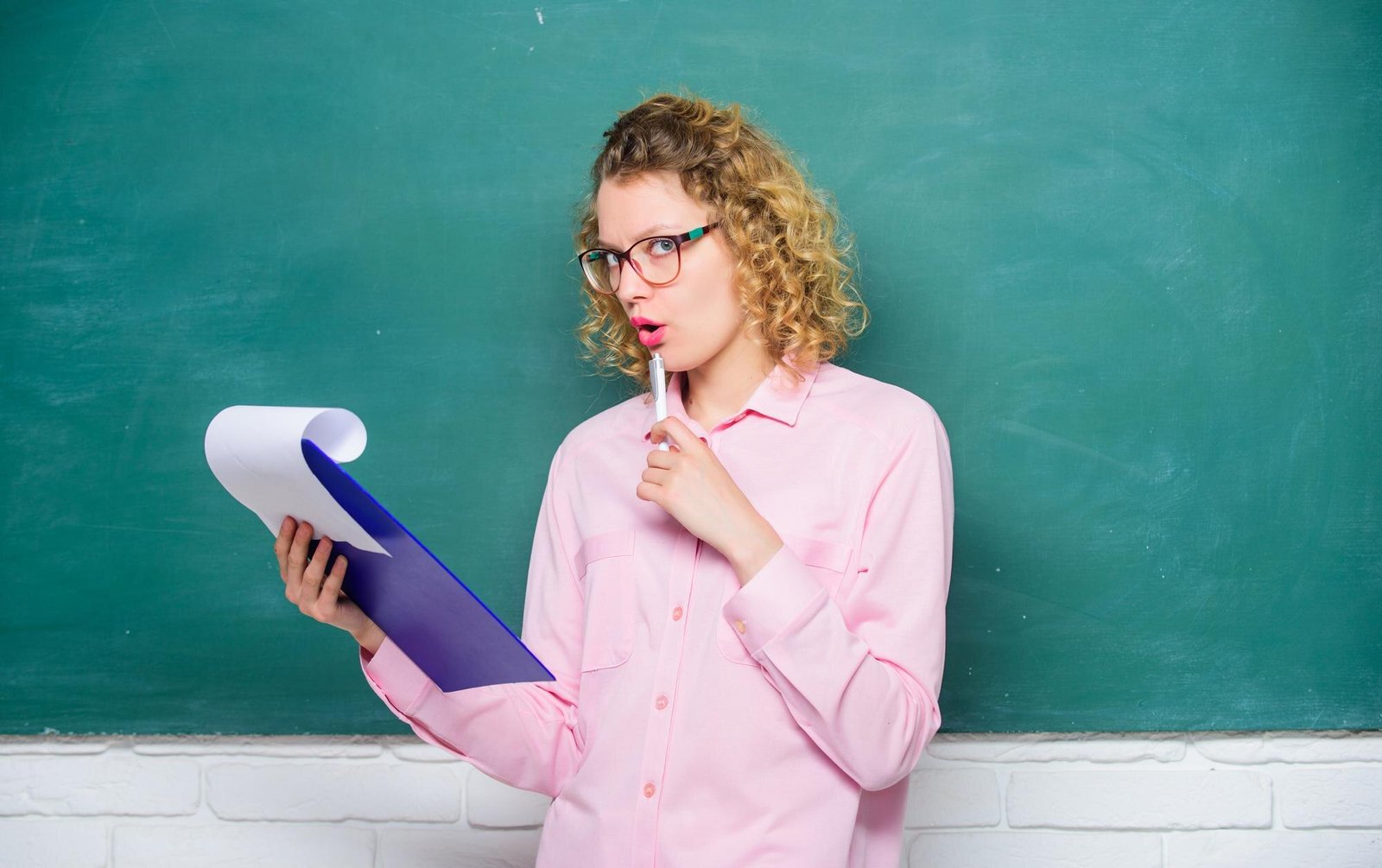 A woman with curly blonde hair and glasses, wearing a pink shirt, holds a large purple and white question mark prop and a pink pen while looking to the side against a teal background.