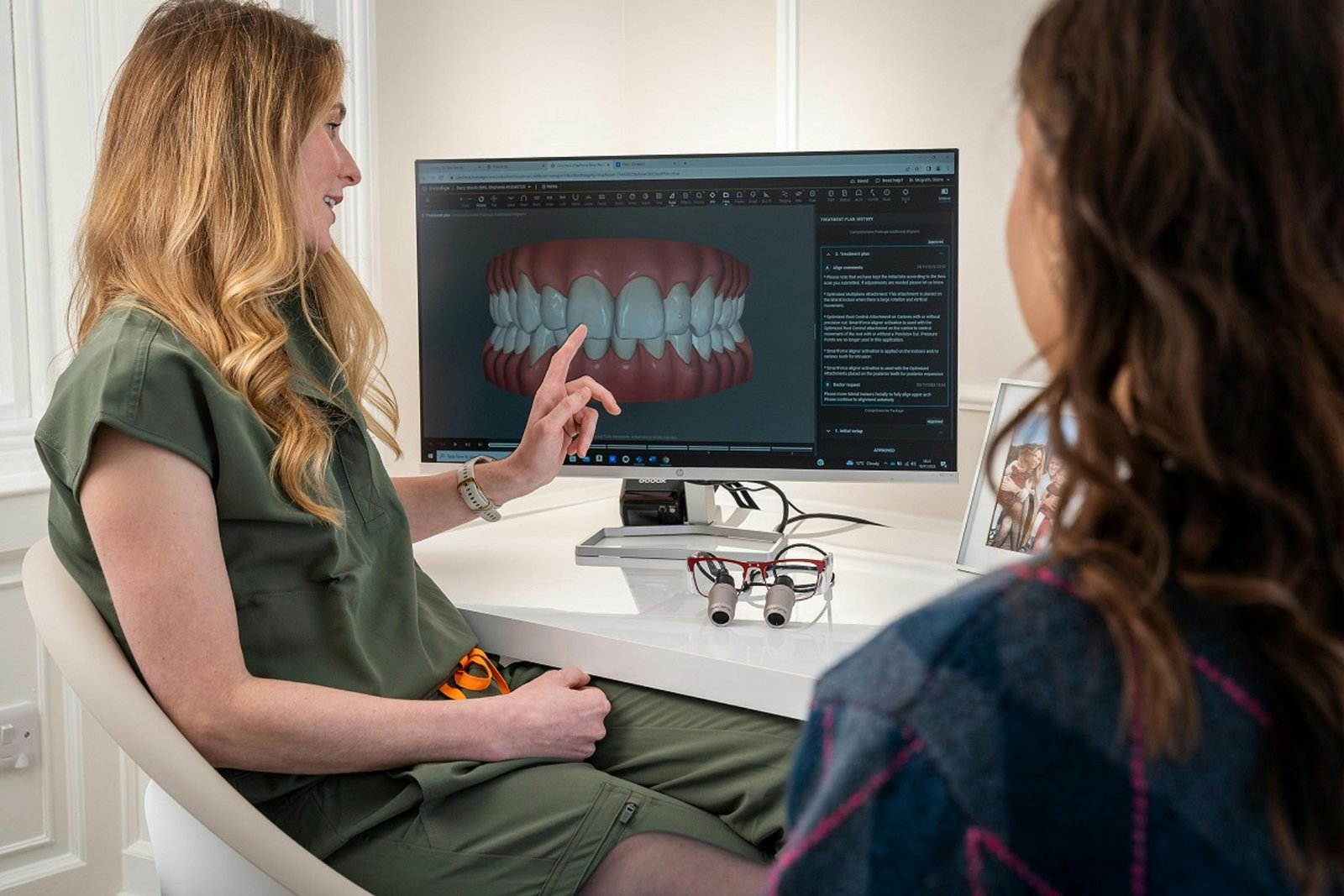 Two women in a contemporary dental education setting view a digital dental imaging display on a computer monitor showing a panoramic X-ray of teeth; the woman on the left wears a green medical uniform and appears to be instructing or discussing the image with the woman seated on the right.