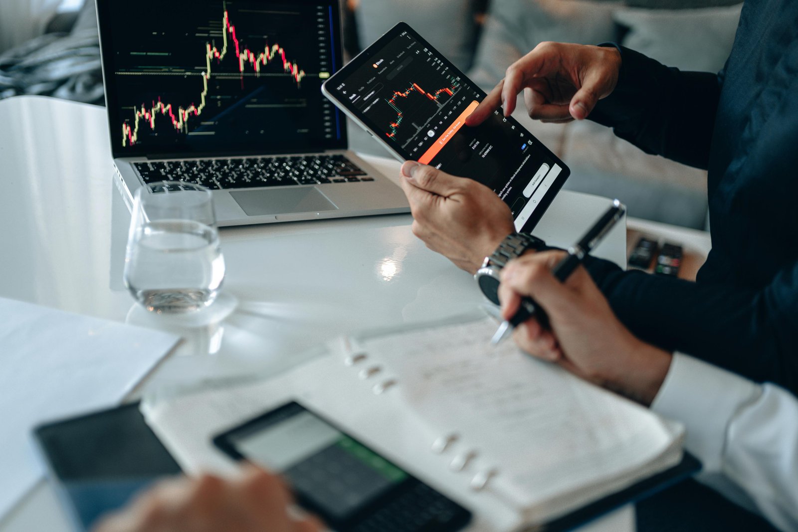 Two business professionals in an office setting examine financial charts on a tablet and laptop displaying candlestick graphs and market data, with a glass of water and documents visible on the desk.