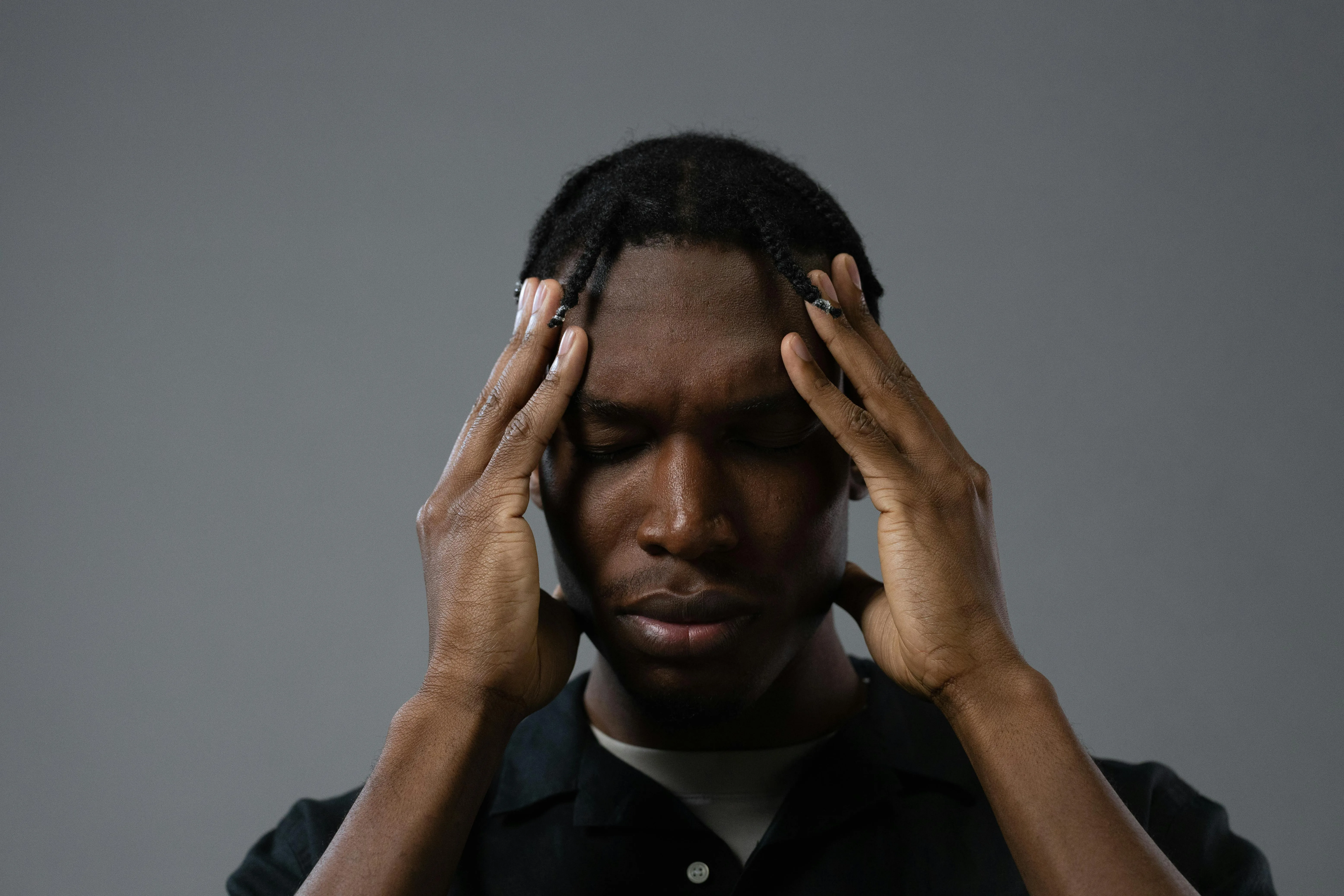 A person with closed eyes and a thoughtful expression, with both hands placed on their head, against a neutral gray background.