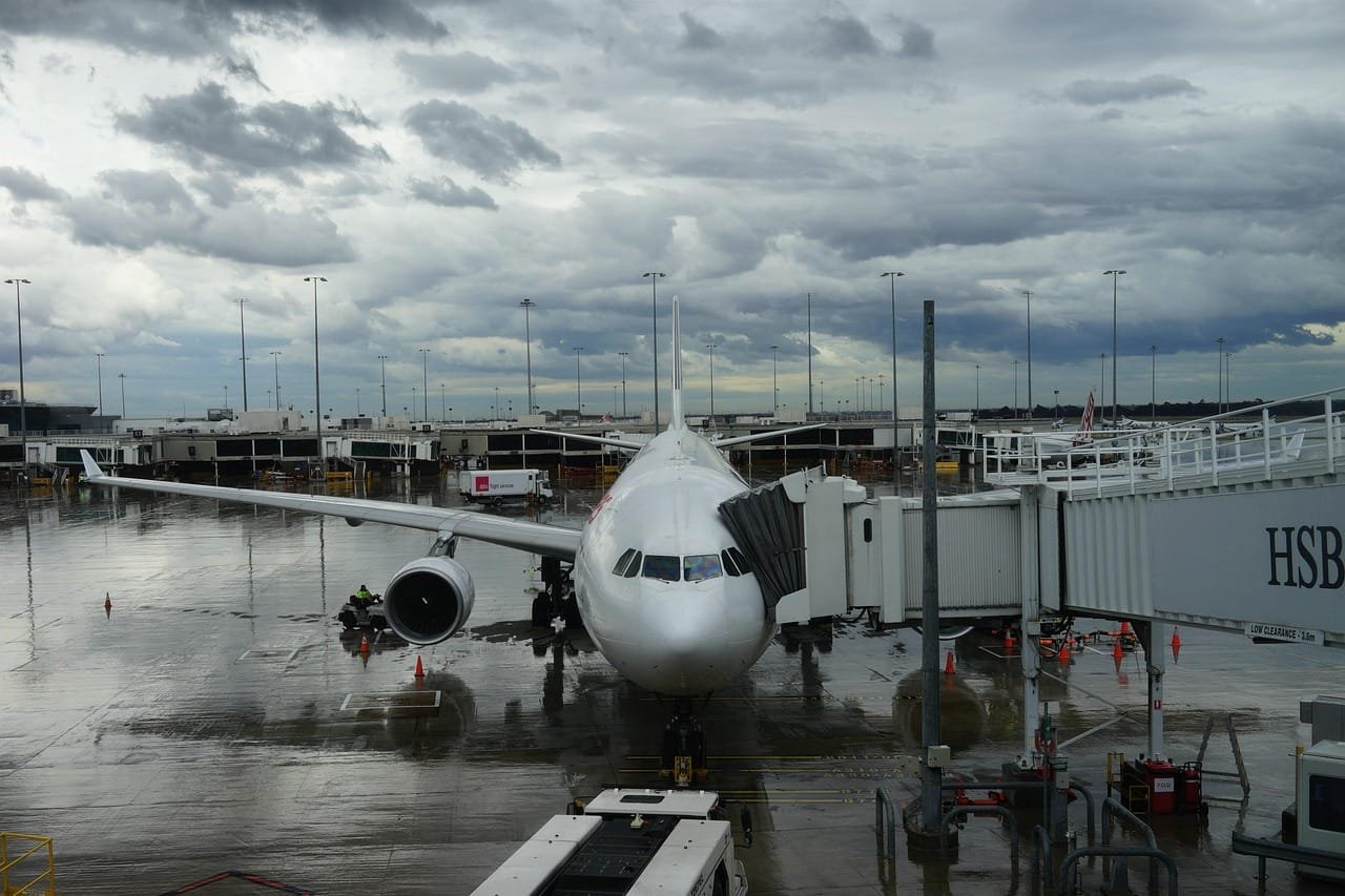 A commercial airliner parked at an airport gate with ground support equipment and airport infrastructure visible under an overcast sky.