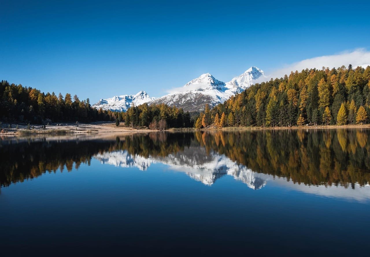 A forested mountain landscape with a calm, mirror-like lake in the foreground reflecting snow-capped peaks and evergreen trees under clear blue sky.