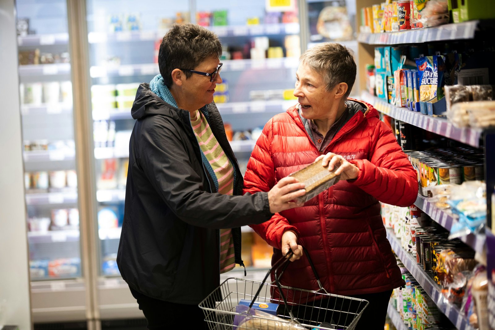 Two adults shopping together in a grocery store, with one person holding a shopping cart while selecting food items from shelves stocked with packaged goods and beverages.